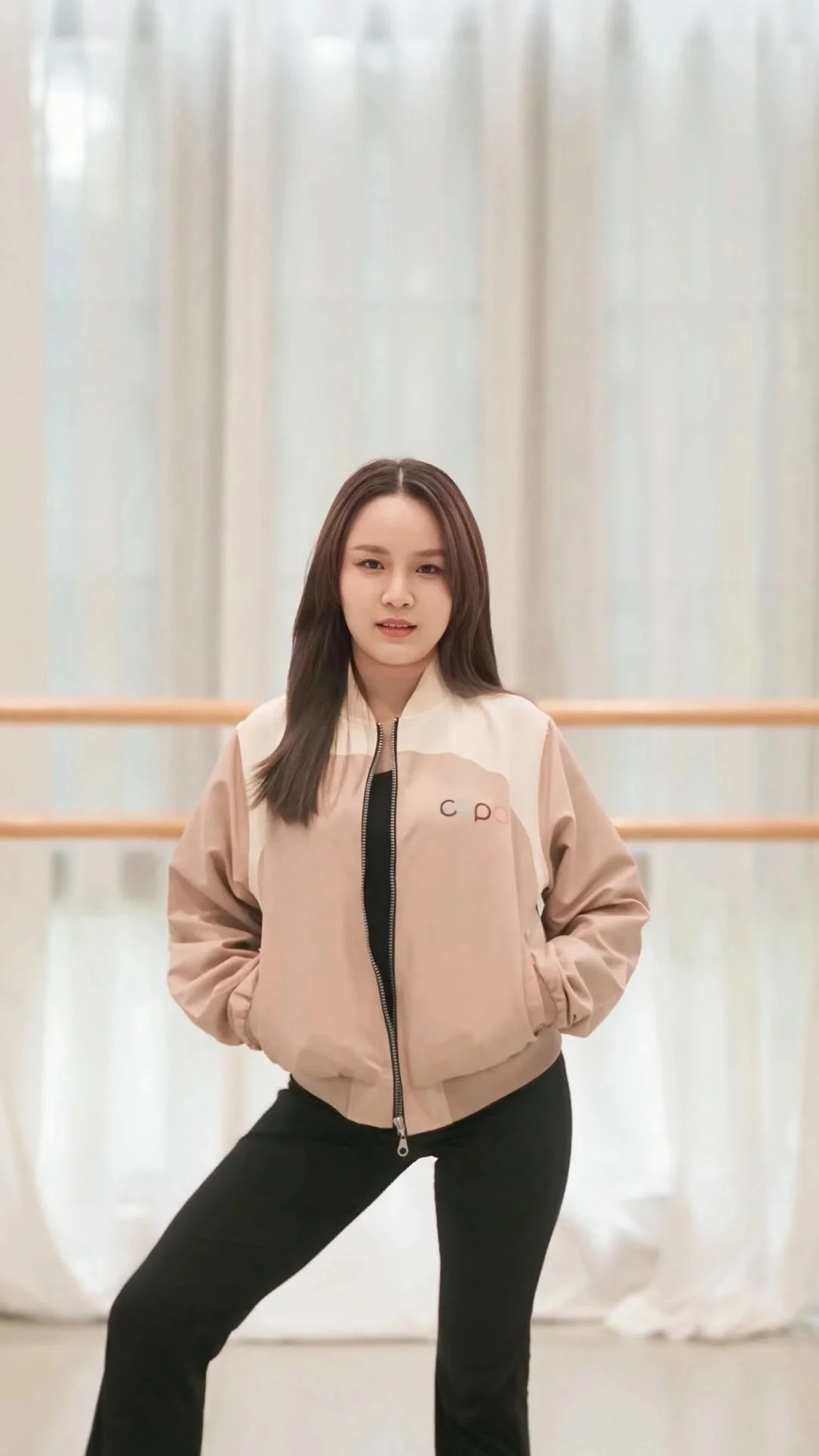 Young woman with shoulder-length brown hair standing in a dance studio, wearing a beige and white zip-up jacket and black pants.
