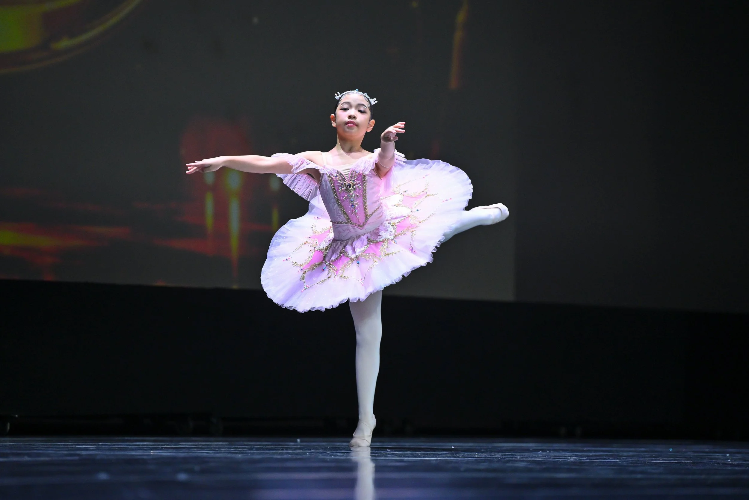 A young ballerina in a pink tutu performing a ballet dance on stage, balancing on one leg with her arms outstretched.