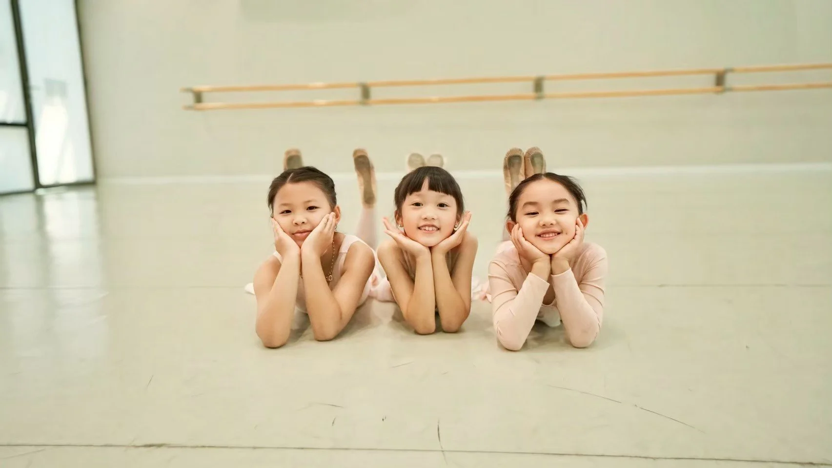 Three young girls lying on their stomachs on a dance studio floor, resting their heads on their hands and smiling at the camera.
