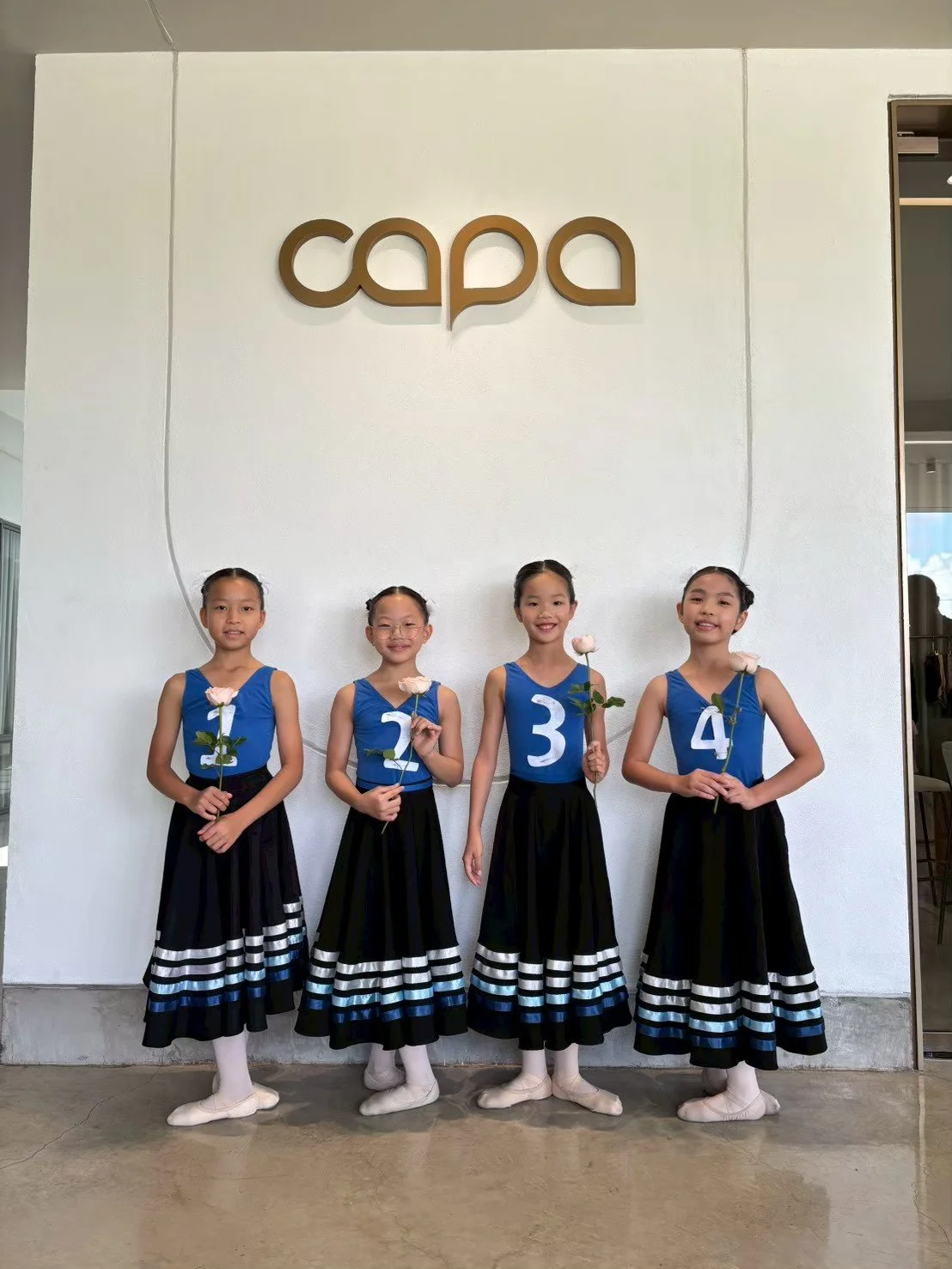 Four young girls in blue and black ballet costumes holding roses, standing in front of a white wall with a wooden sign that reads 'capa'.