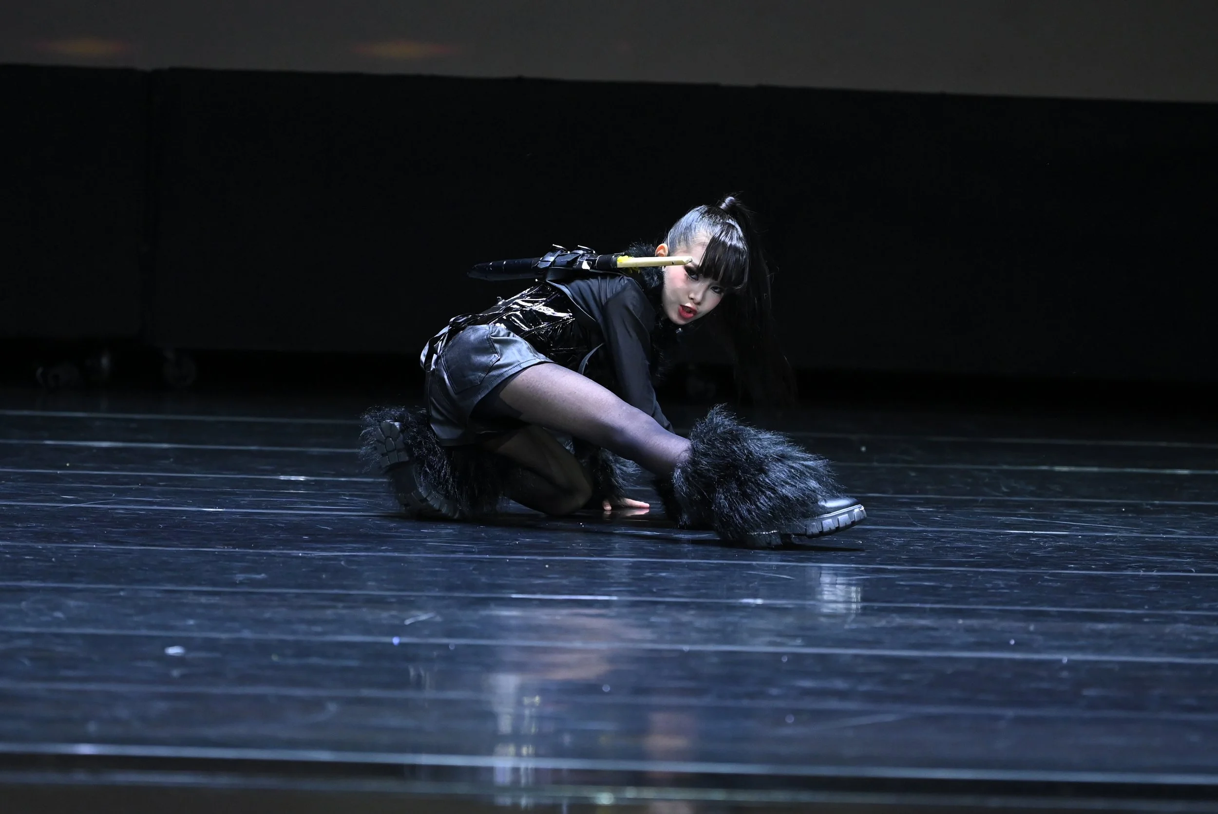 A young woman on stage dressed in black with wolf-like accessories, crouching on the stage floor in a dramatic pose during a performance.