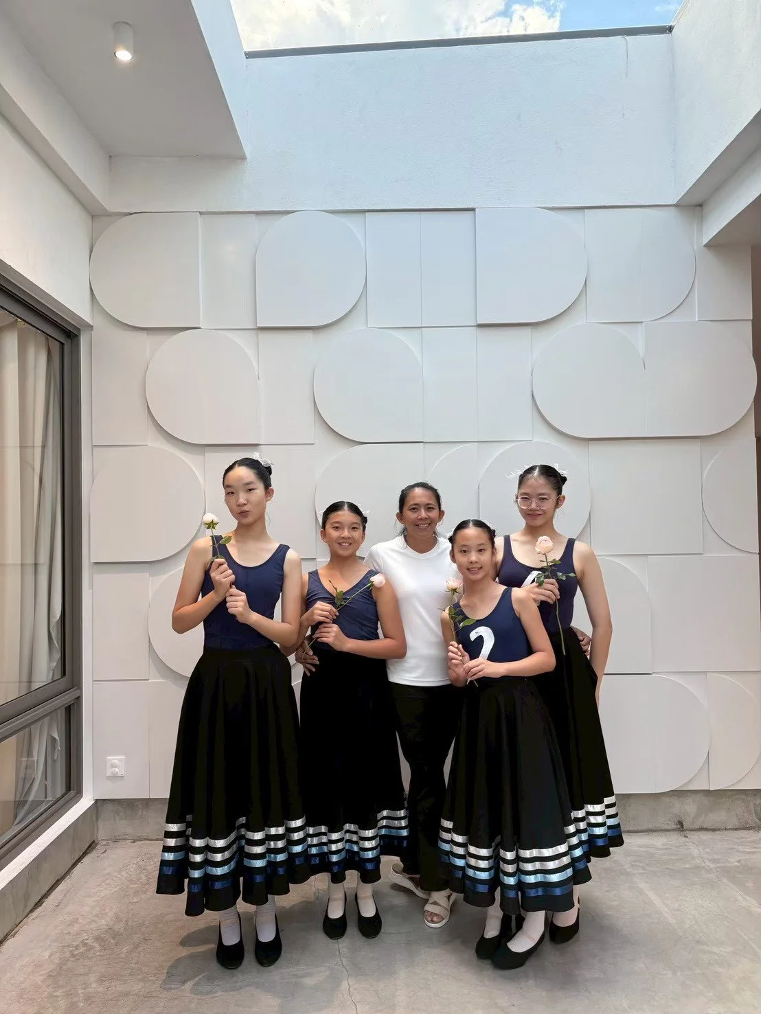 A group of five girls and one woman standing inside in front of a white textured wall. The girls are dressed in matching navy blue tops and long black skirts with white and blue stripes near the hem, holding white roses. The woman in the center is we