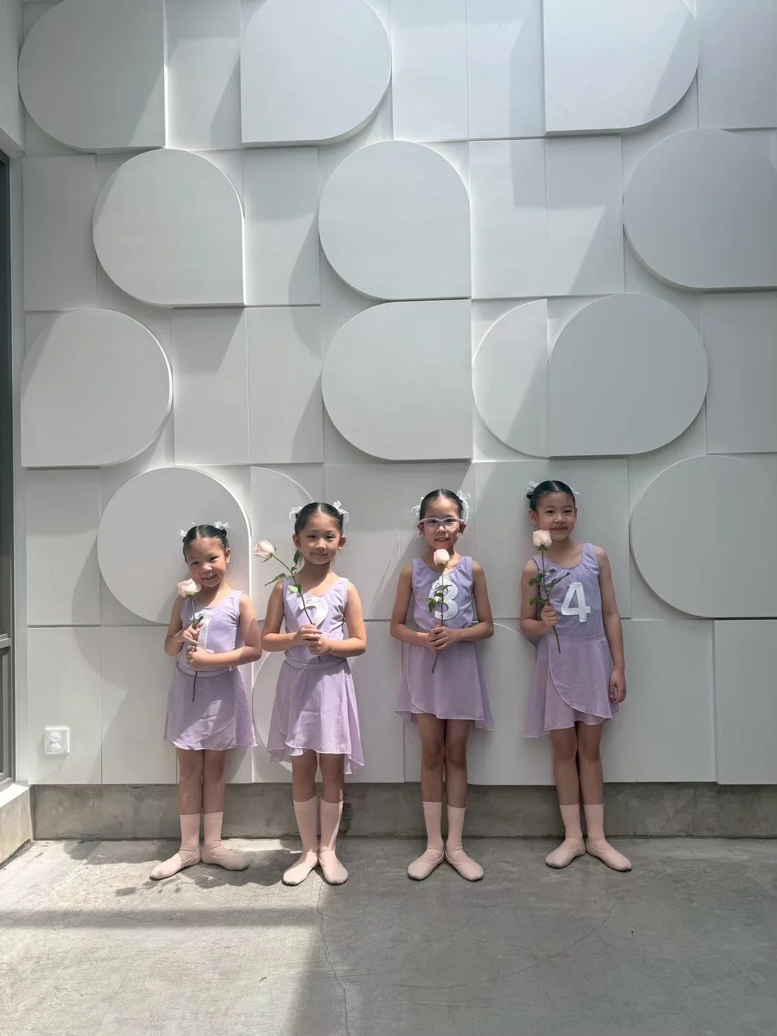 Four young girls in lavender dresses holding pink roses, standing in front of a white geometric wall. They are smiling and wearing socks and ballet slippers, with some girls having hair accessories. Each girl holds a flower and has a number on her dr