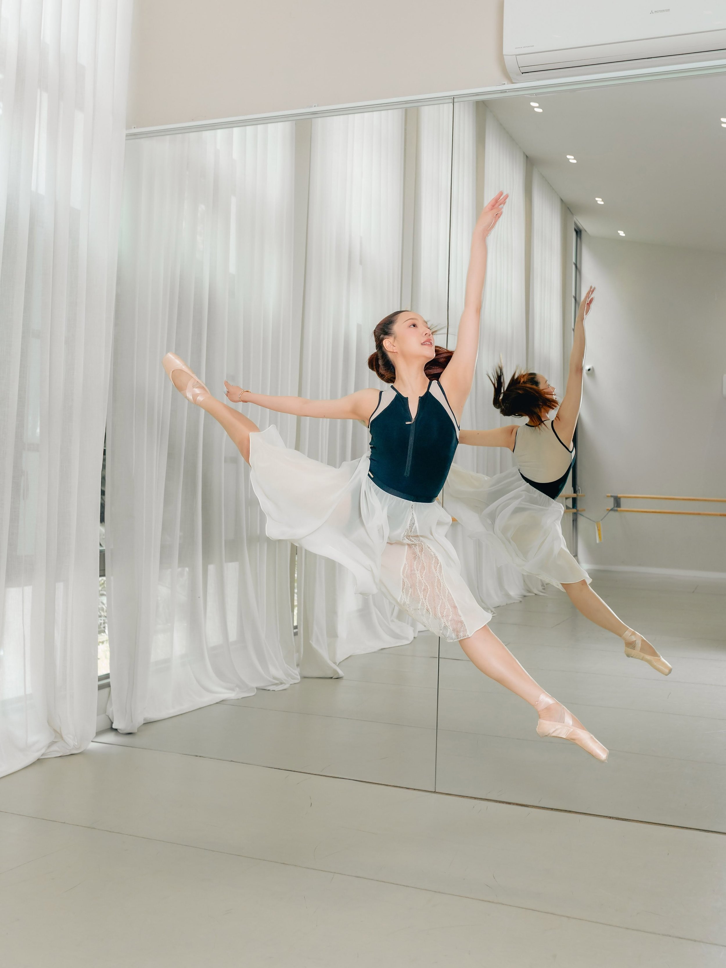 Two ballet dancers practicing in a dance studio with large mirrors and white curtains, one dancer extending her leg and arms in a graceful pose.