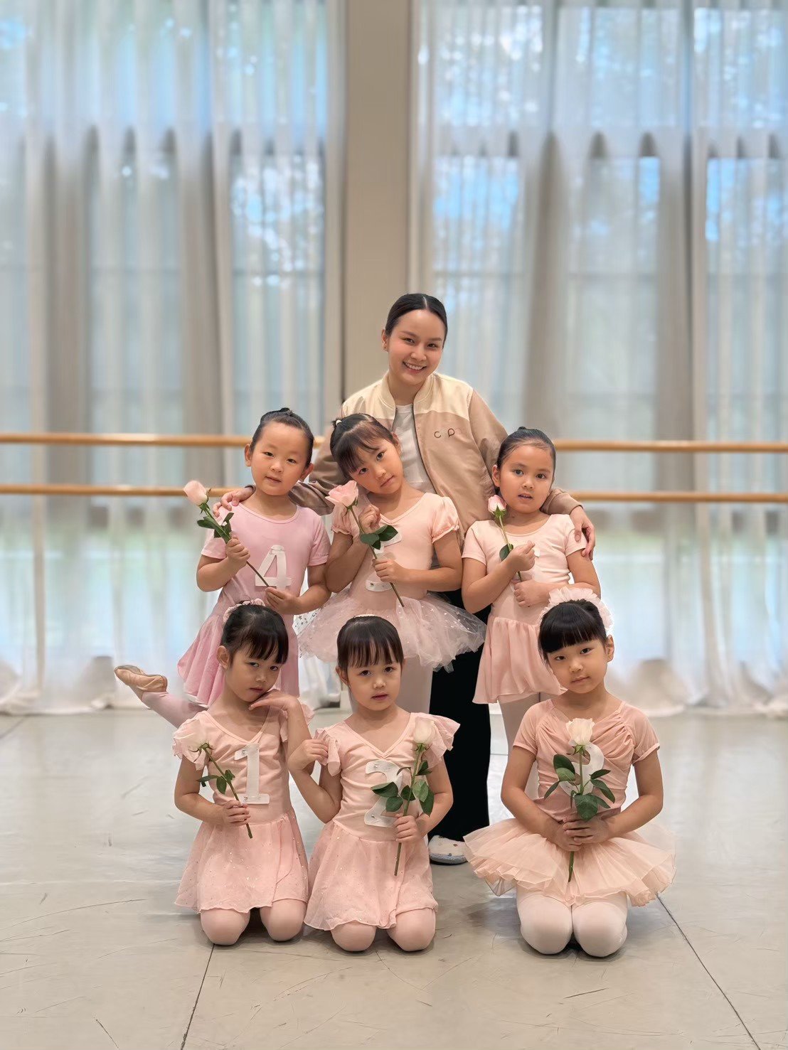 A ballet instructor with seven young girls in ballet costumes holding pink roses. The girls are posing in a dance studio with large windows and ballet barres.