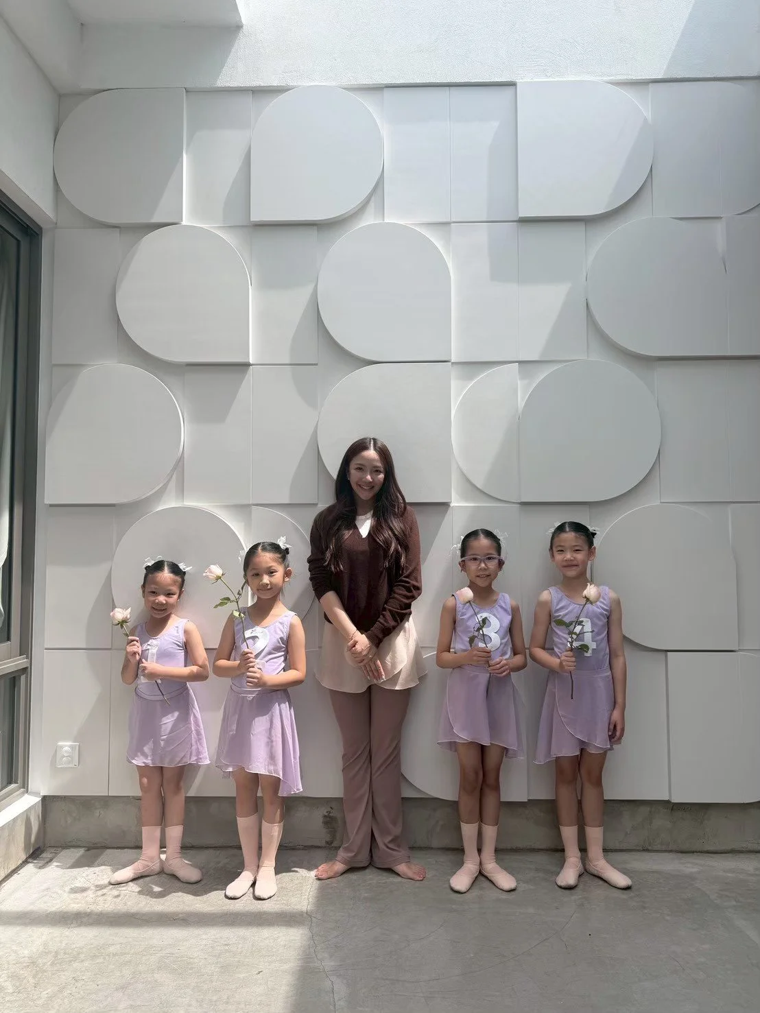 A woman and four young girls in ballet costumes holding flowers stand in front of a geometric white wall.