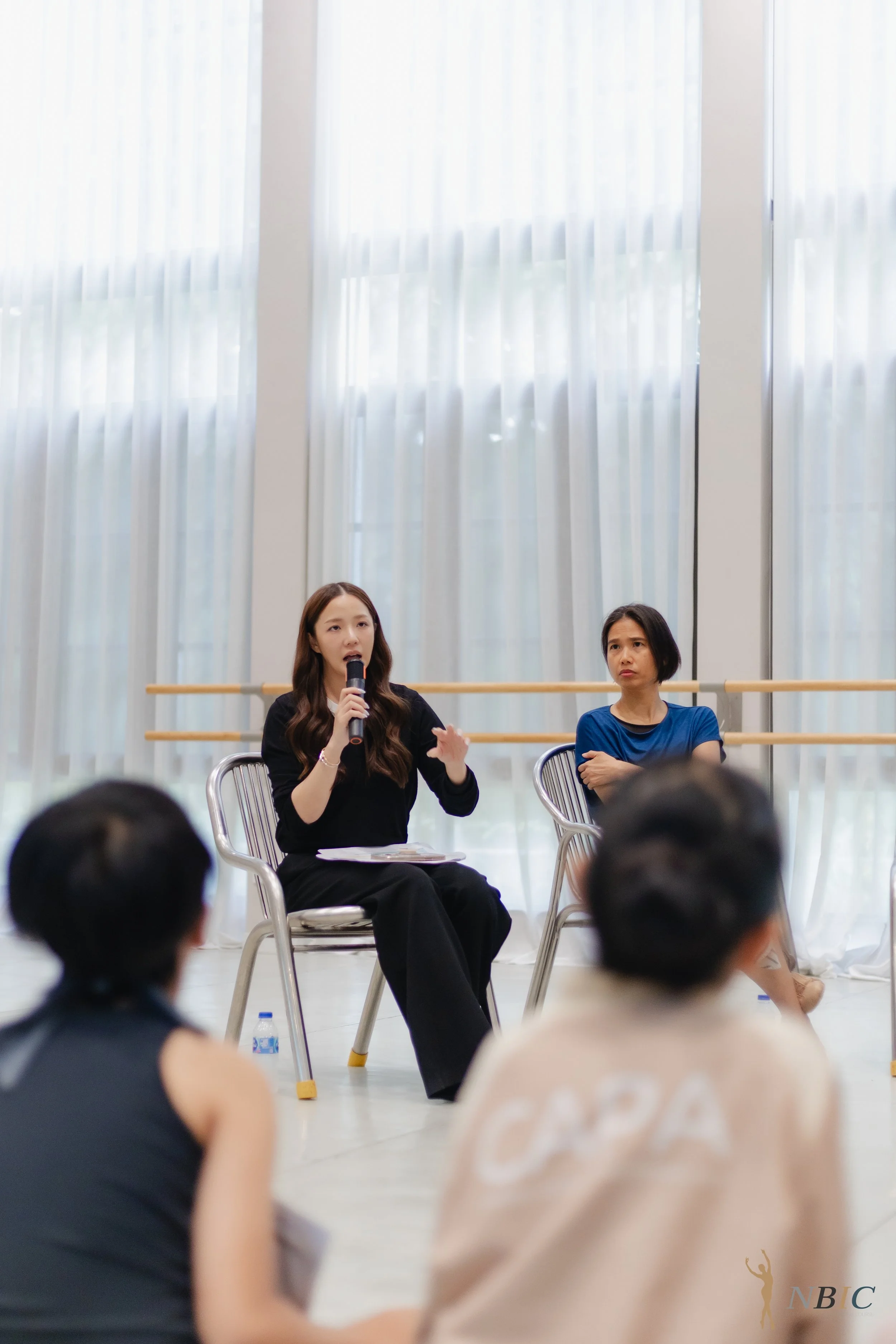 A woman in black clothing holding a microphone and speaking during a panel discussion, seated in a transparent chair. Next to her is another woman in a blue shirt. An audience is visible in the foreground, with heads and shoulders showing, in a brigh