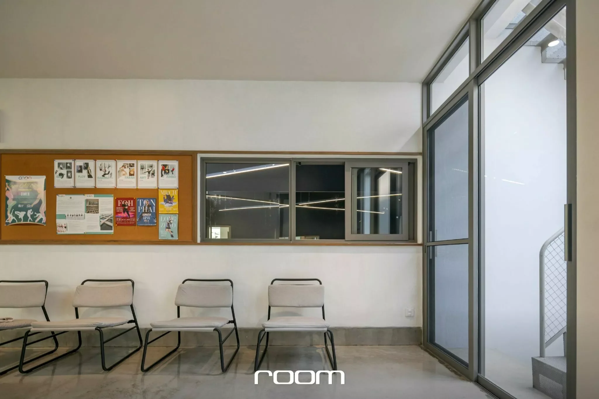 Interior of a waiting area with five white chairs against a white wall, a cork bulletin board with posters, a glass window, and a glass door with a metal frame.