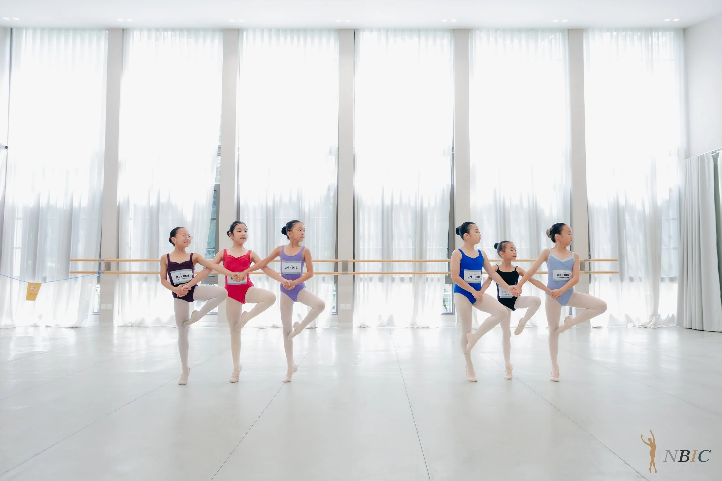 Six young girls practicing ballet in a dance studio with large windows and sheer curtains. They are wearing colorful leotards and tights, holding hands in a circle with one leg raised. A small logo of a dancer silhouette with the letters 'NBIC' is vi
