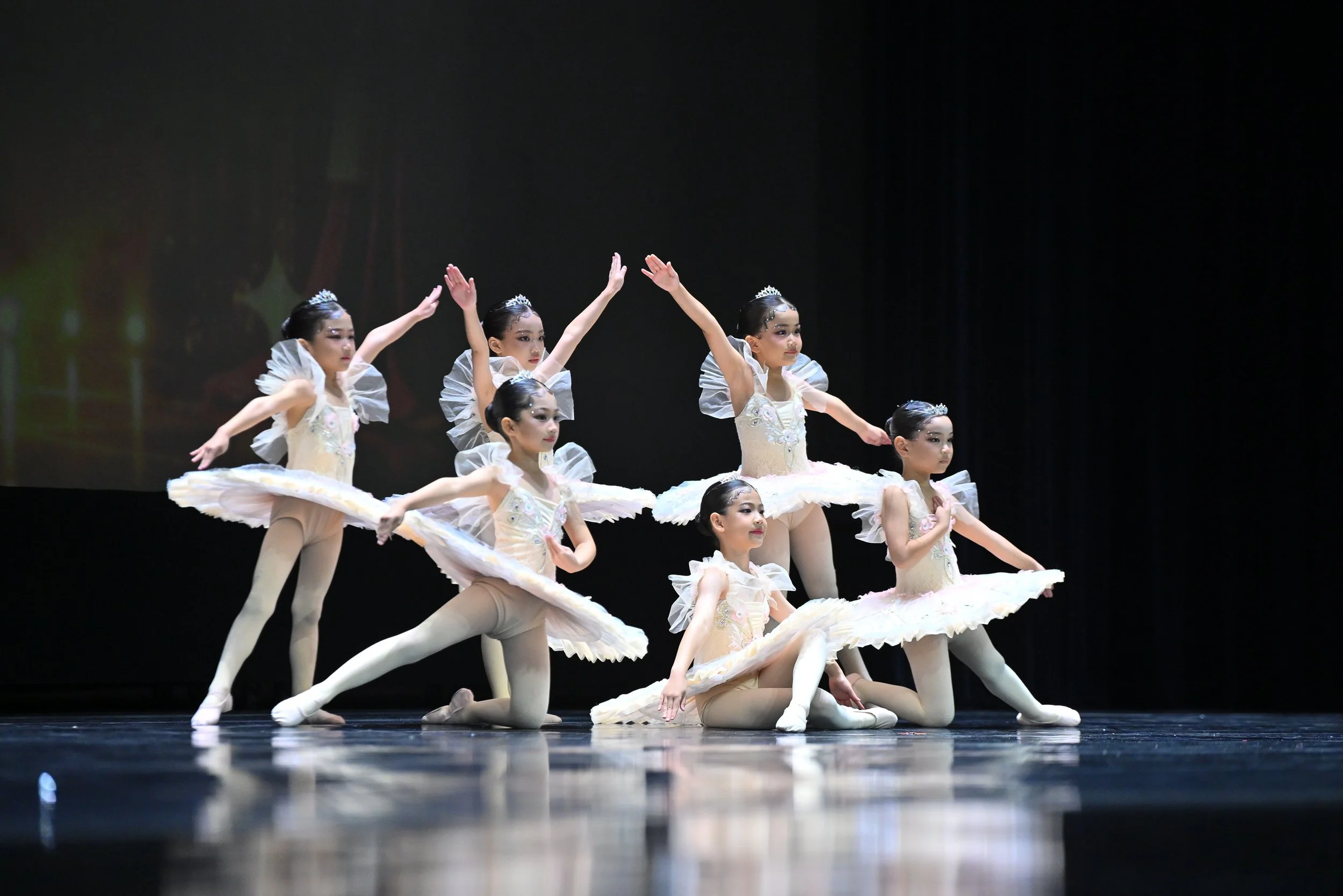 Group of young ballerinas in cream-colored tutus performing a dance on stage, with some dancers kneeling and others standing, wearing tiaras and makeup.