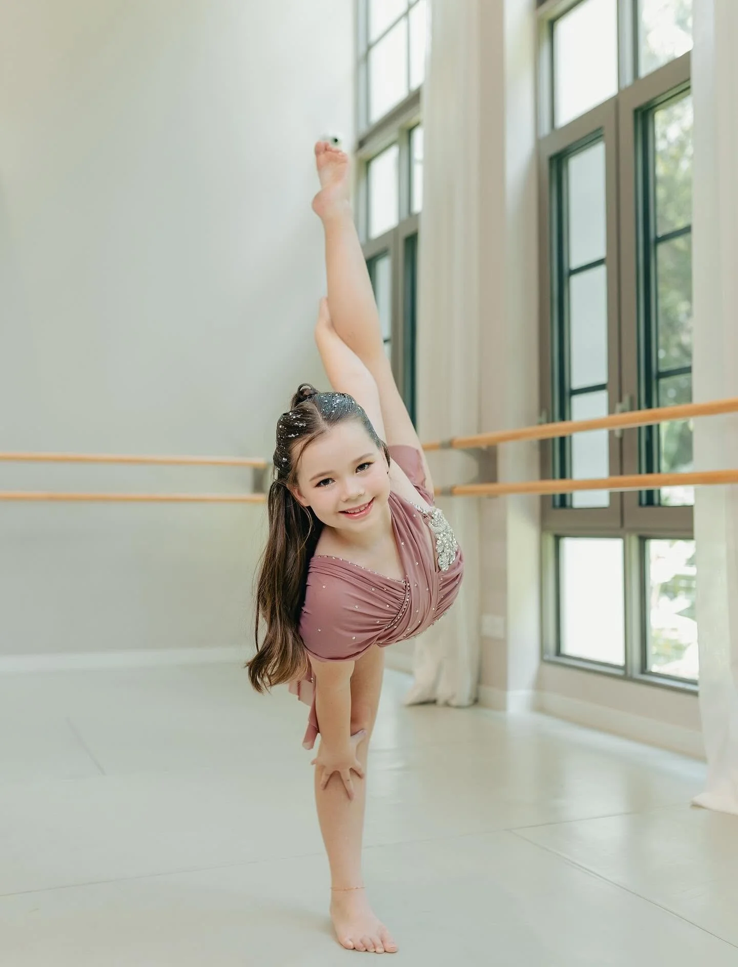 Young girl in ballet attire practicing dance in a studio with large windows and wooden ballet barre.