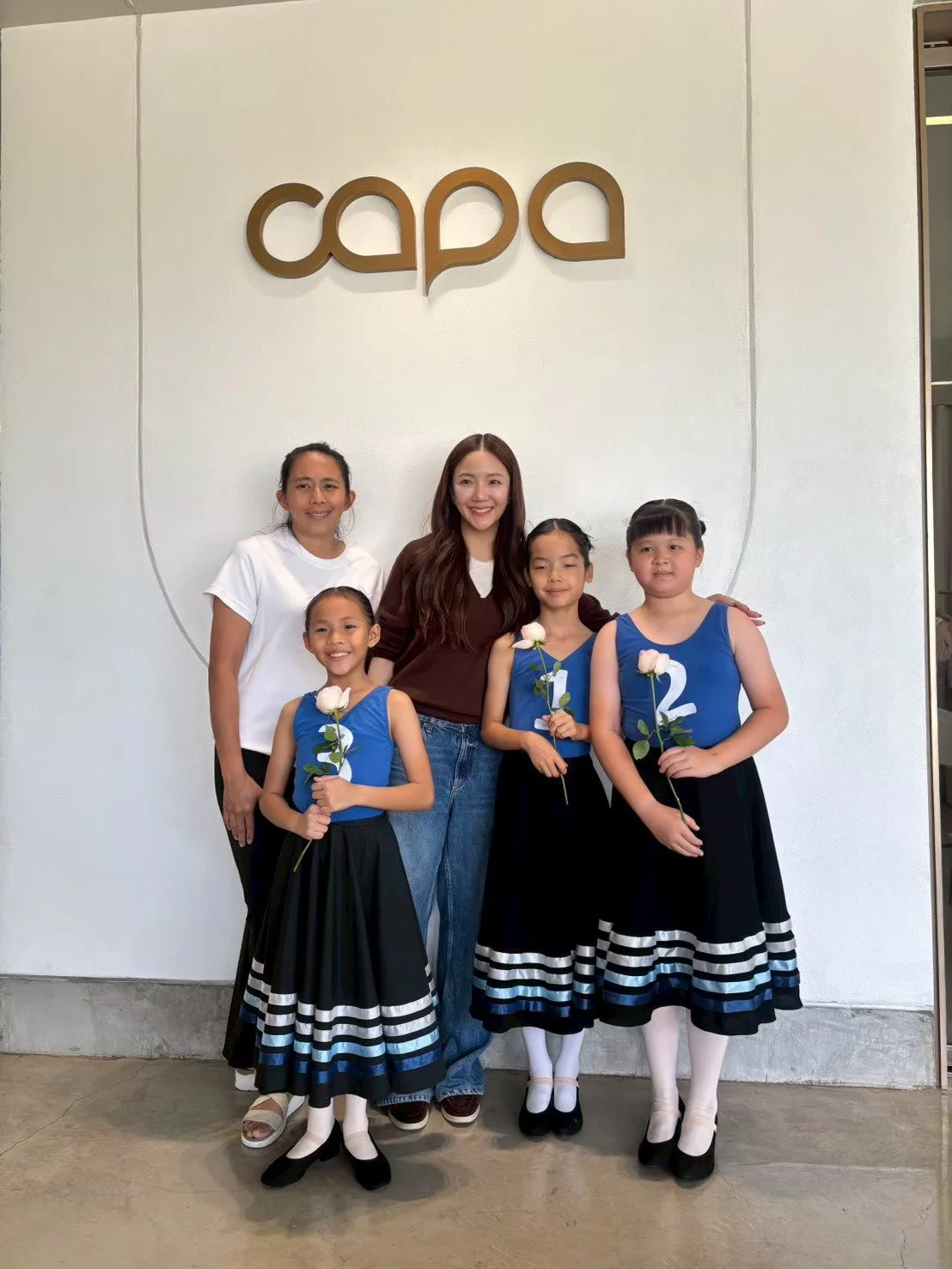 Group of five females standing in front of a white wall with a logo that says "capa." The three young girls are dressed in blue dance costumes with black and white striped skirts, and each is holding a white rose. The other two women are standing beh