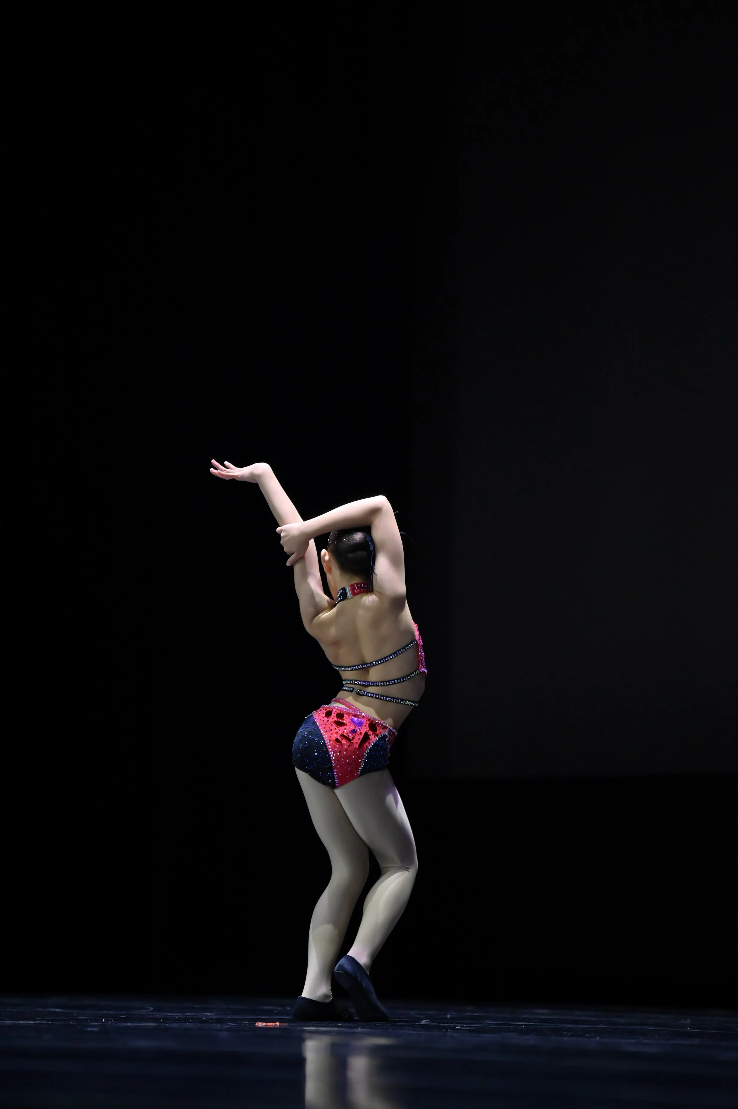 A female gymnast performing a dance move on stage, dressed in a colorful, sparkly leotard with a black background.