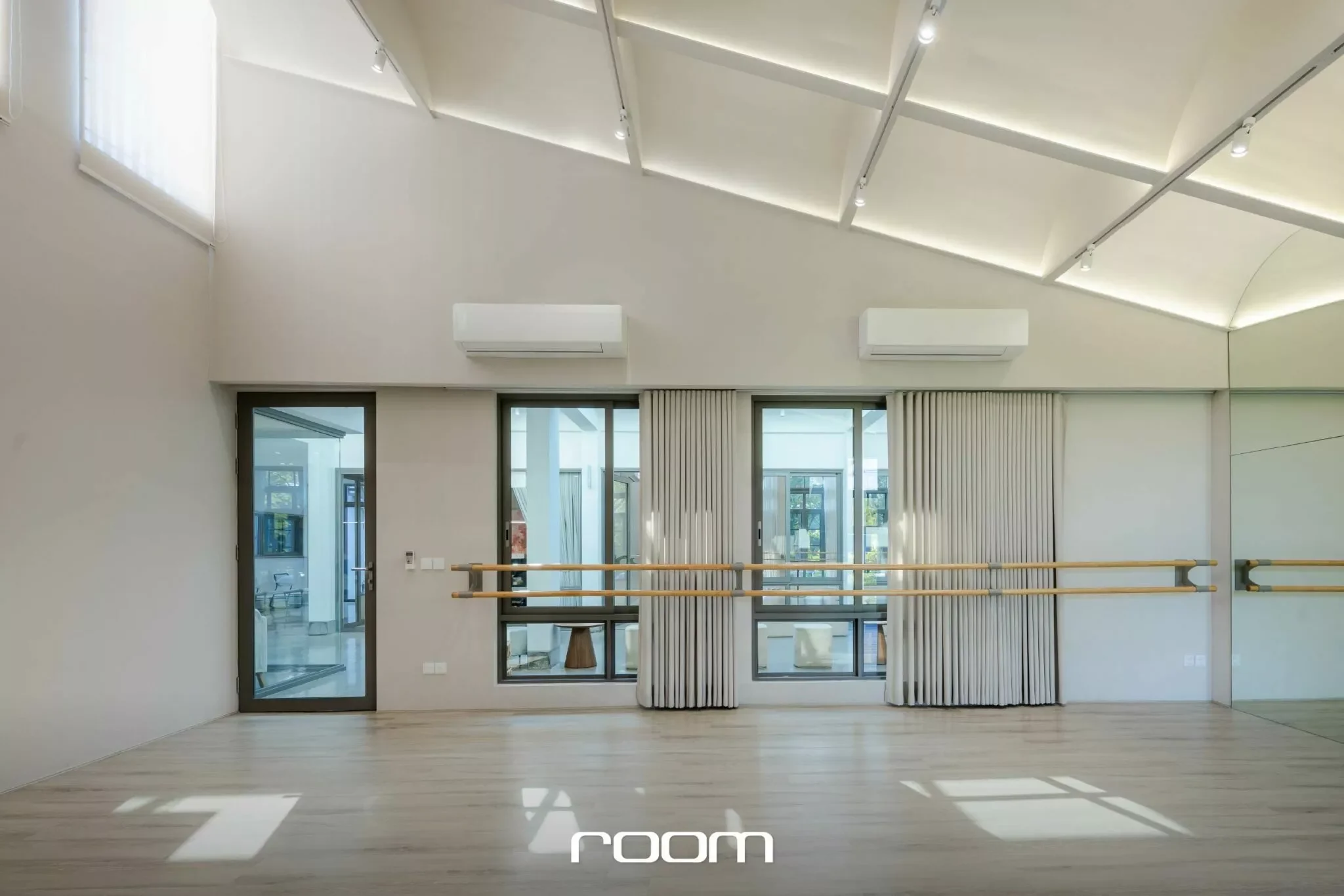 Empty dance studio with large windows, wooden floor, and ballet barre. Modern ceiling with adjustable lighting. Curtain partially closed over window.