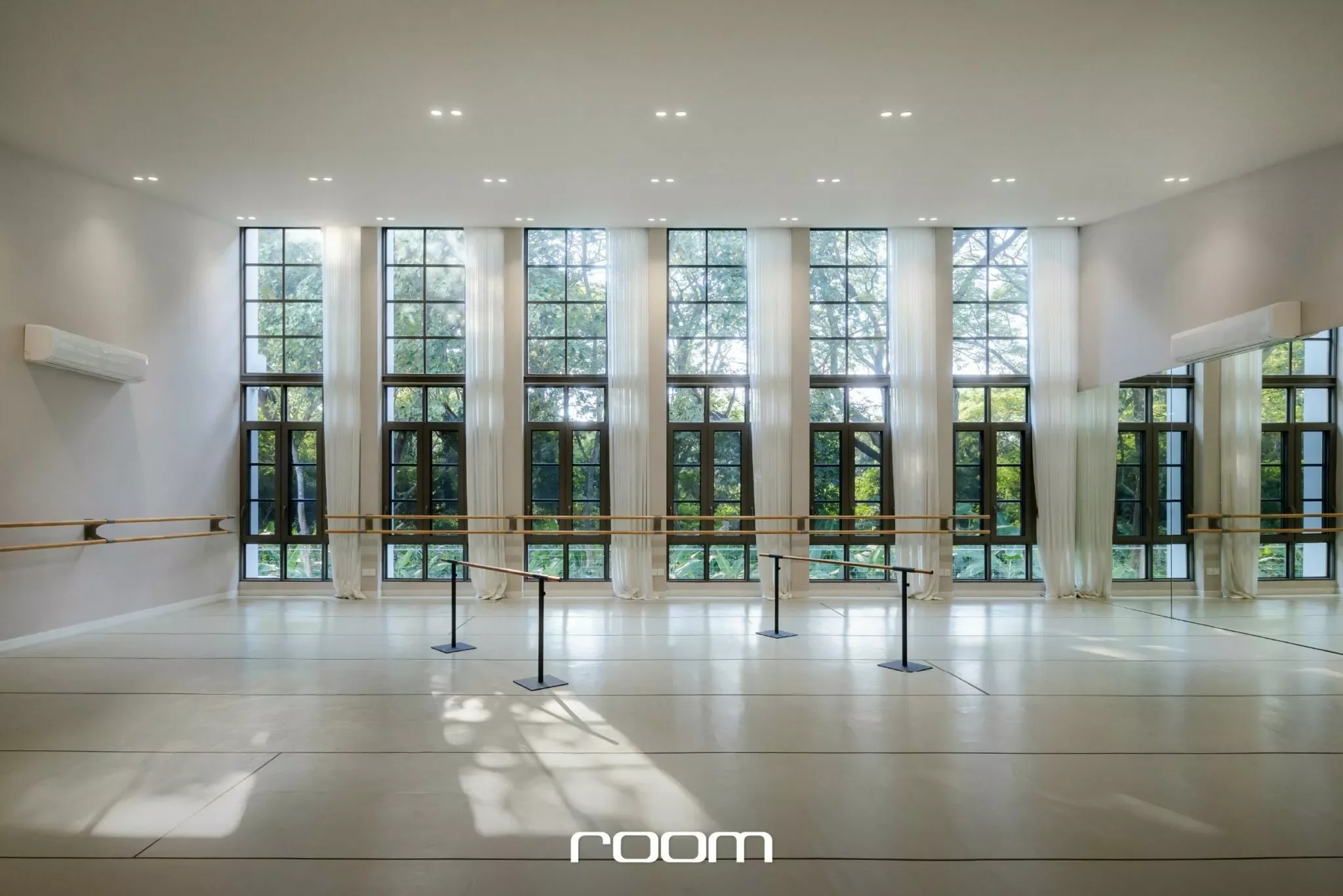 Empty dance studio with large windows showing trees outside, ballet barres along the walls, and soft natural light.