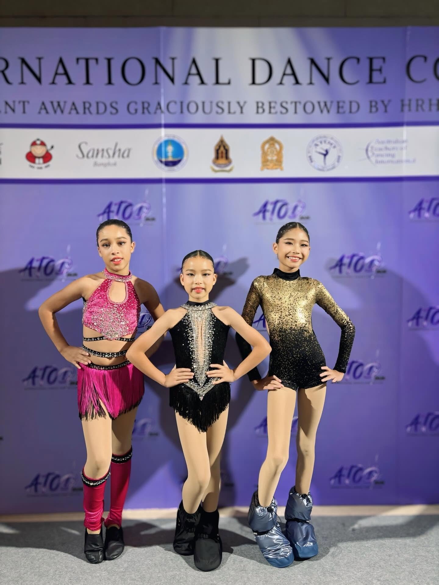 Three young female figure skaters in competition costumes posing with hands on hips in front of a purple backdrop with text and logos.