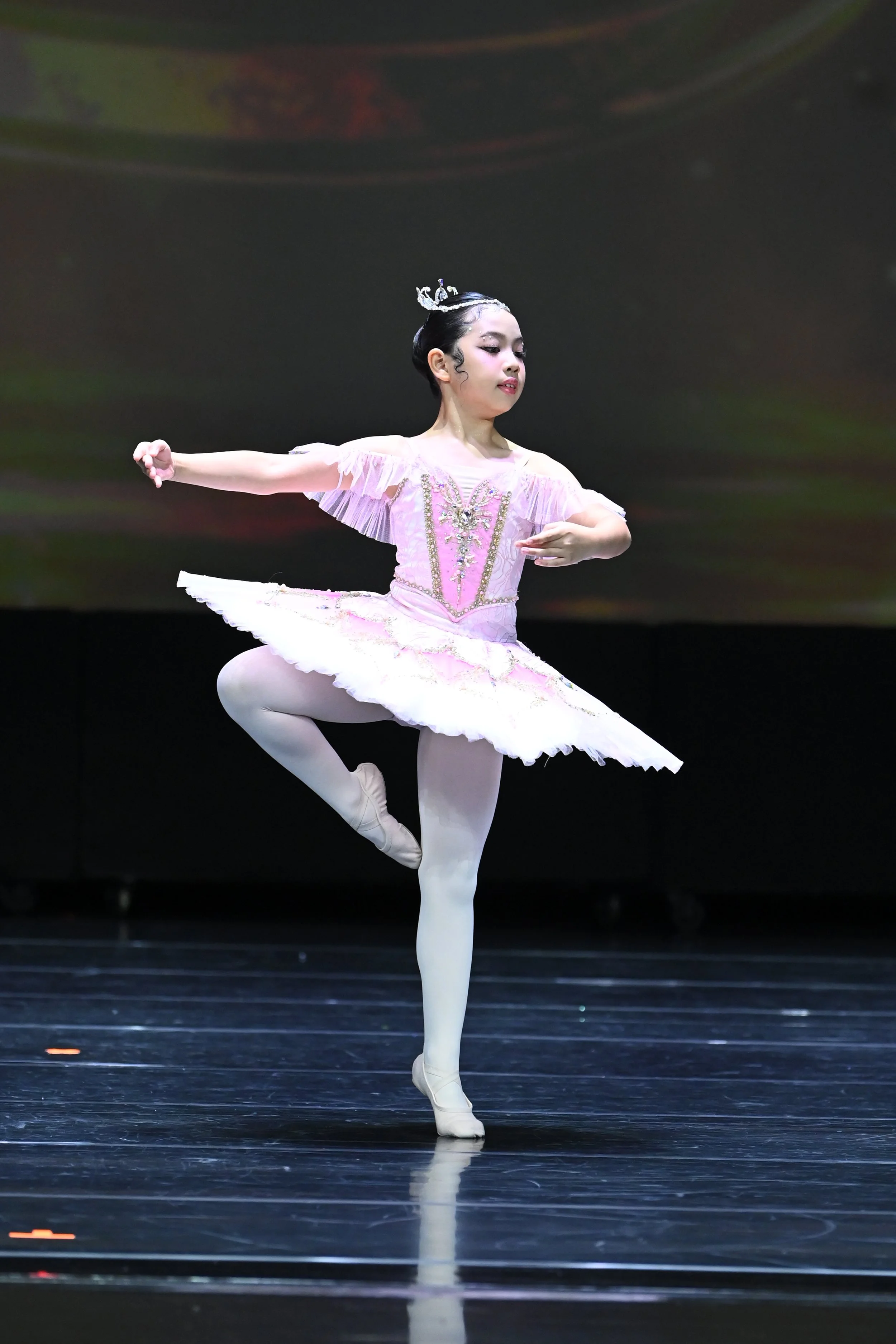 A young ballerina in a pink tutu performing a ballet pose on stage with a dark background.