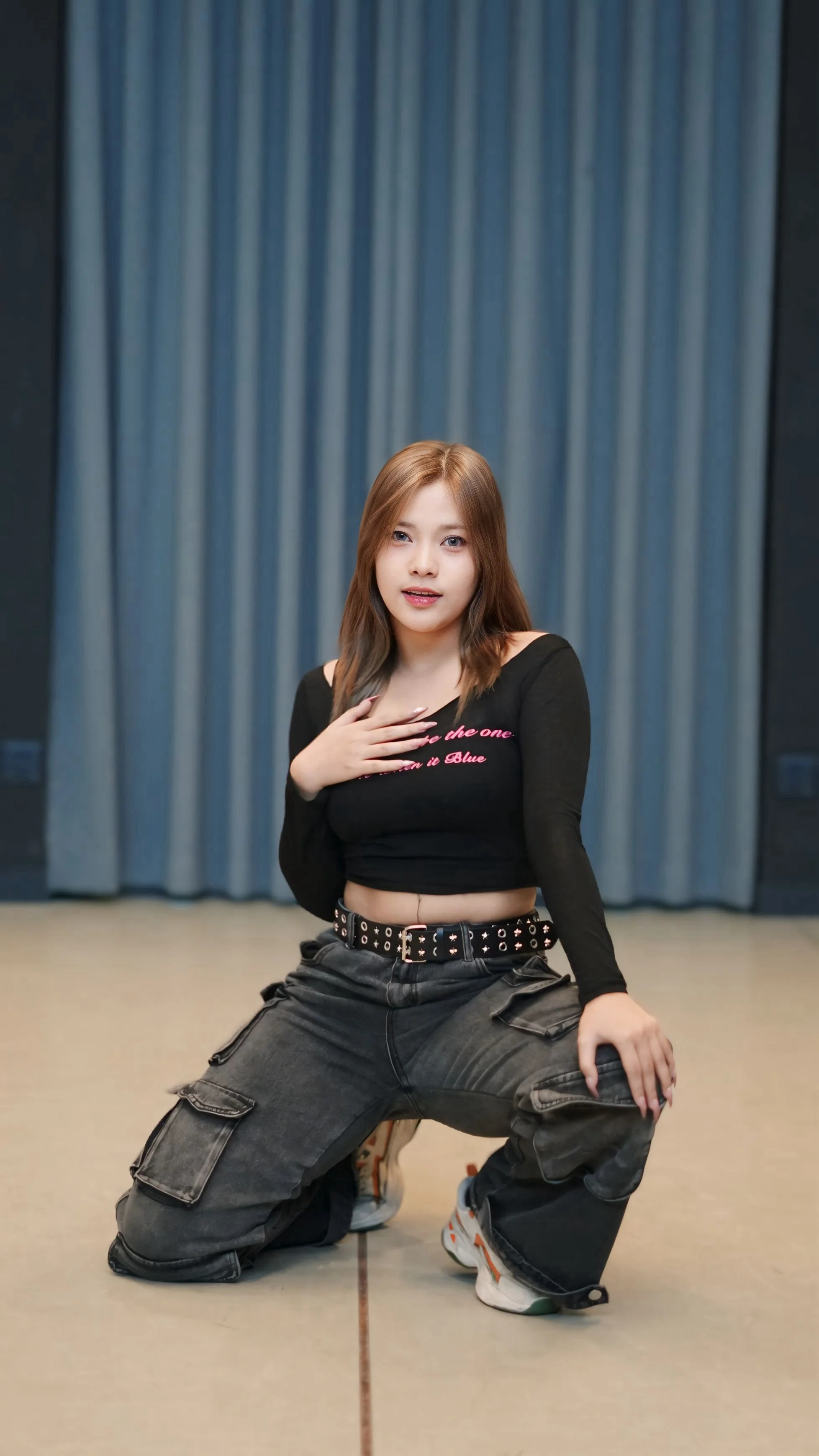 A young woman with light brown hair and fair skin posing with one hand on her chest and the other resting on her knee in front of blue curtains, wearing a black long-sleeve crop top and dark cargo pants.