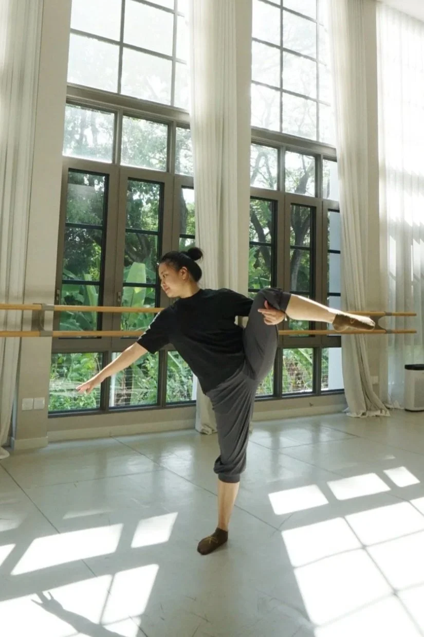 A woman practicing ballet in a bright studio with large windows, dressed in comfortable casual clothes, balancing on one leg with arms extended.
