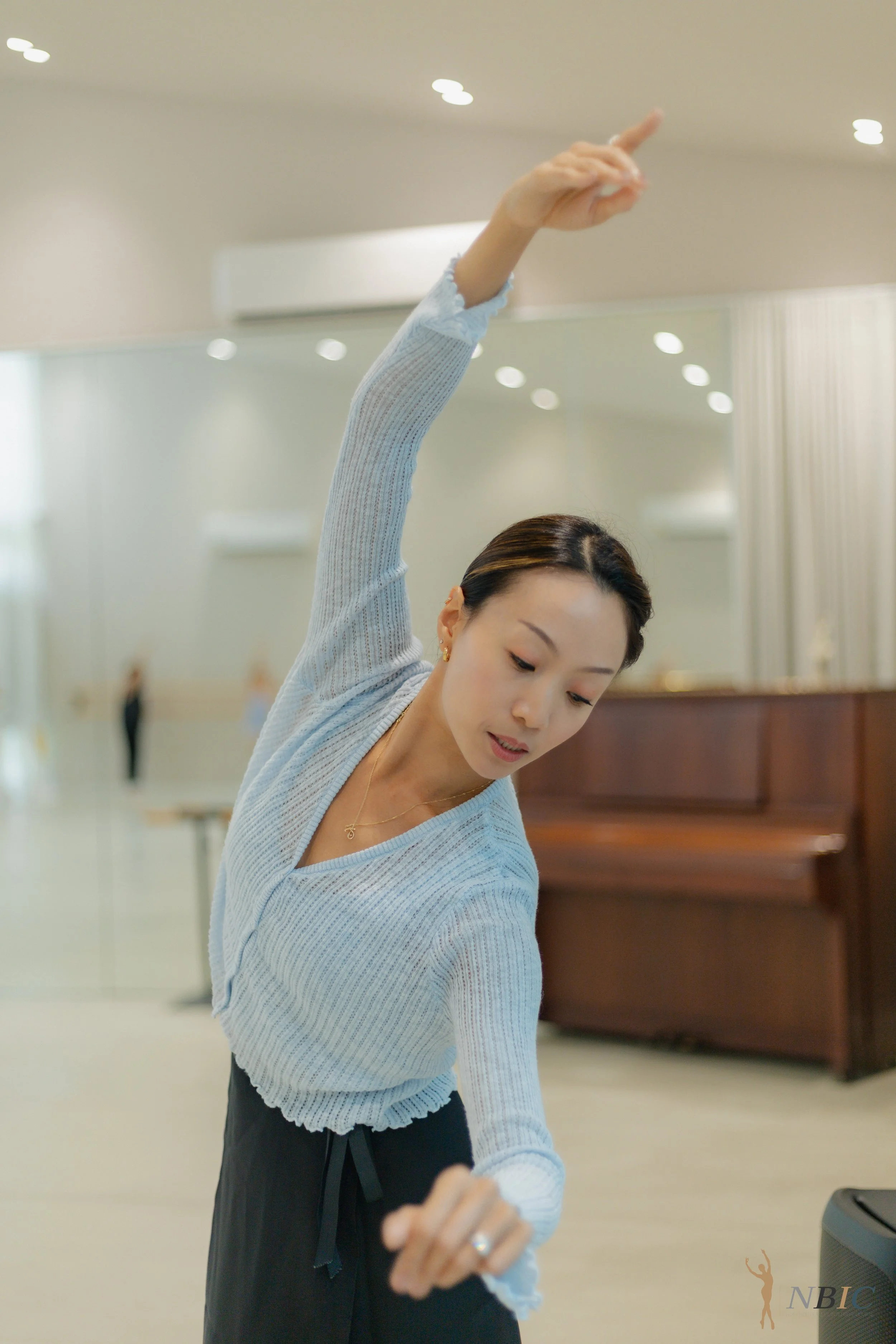 A woman in a light blue sweater and black pants with a drawstring waist is performing a dance move in a dance studio. She has dark brown hair tied back and is looking downward with her arm raised, pointing her finger upward. The studio has a clean, l