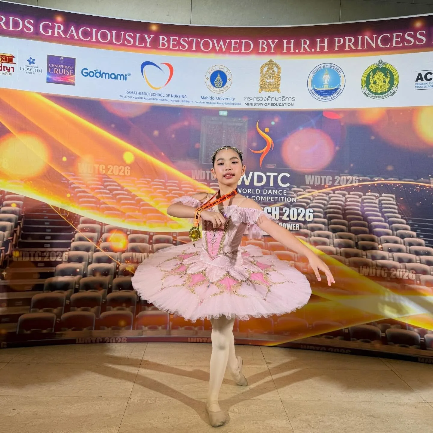A young girl in a pink ballet costume performing a dance in front of a backdrop for a world dance competition, wearing a medal around her neck.