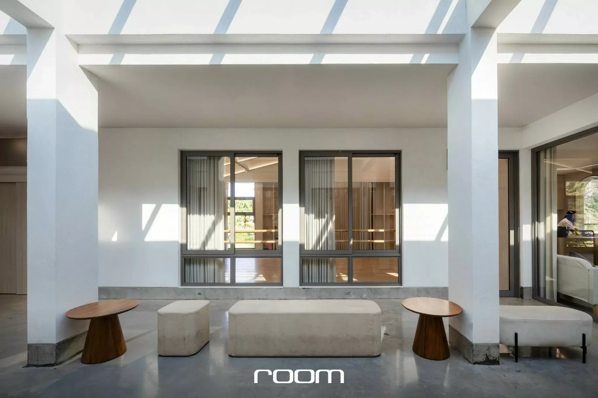 Modern indoor residential space with large windows, white walls, and minimal furniture including round wooden tables and benches.
