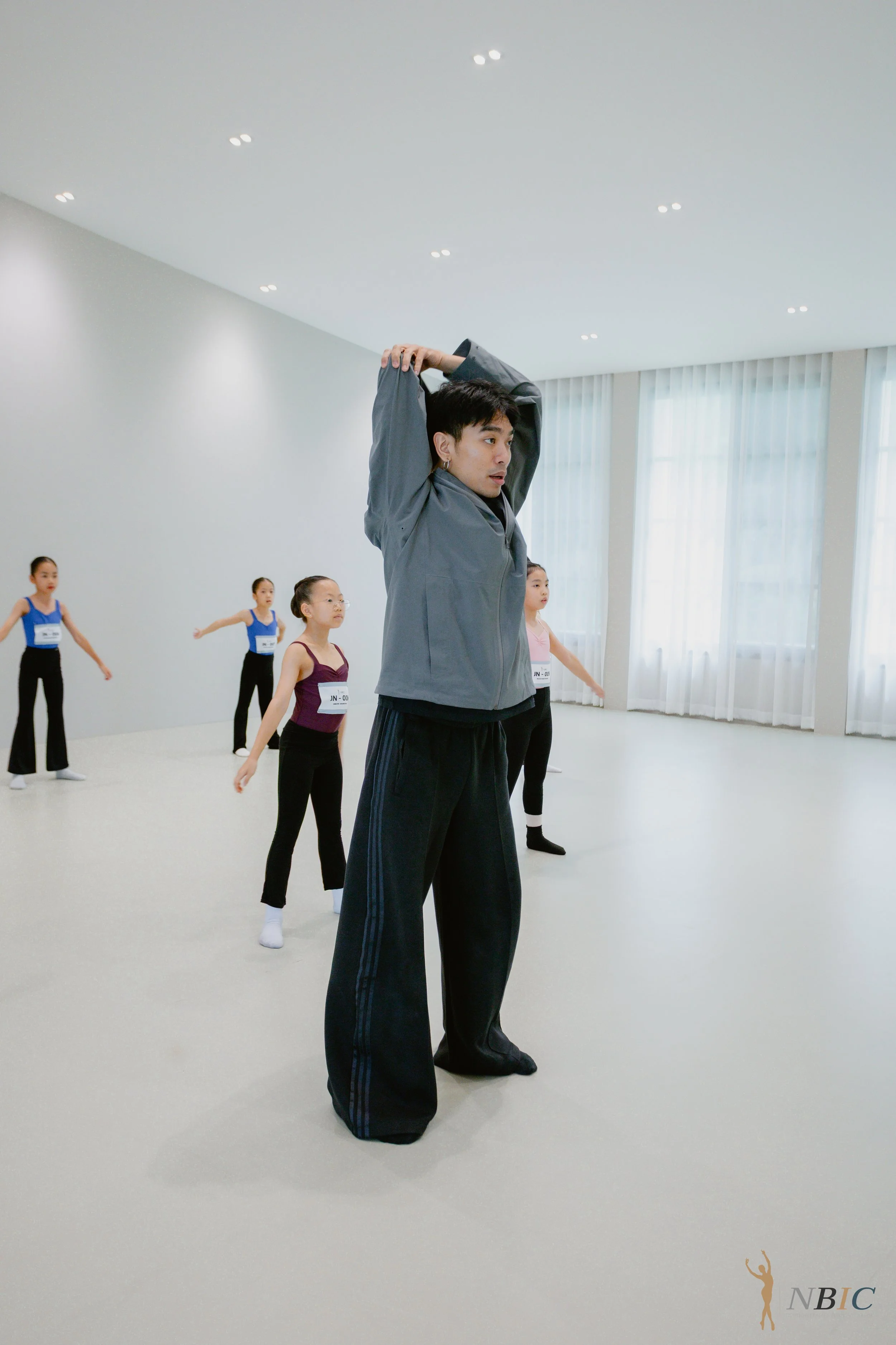 A dance instructor leading a ballet class with young students in a spacious, well-lit studio with white walls and large windows, as the instructor stretches with arms raised overhead and the students stand in a formation behind him.