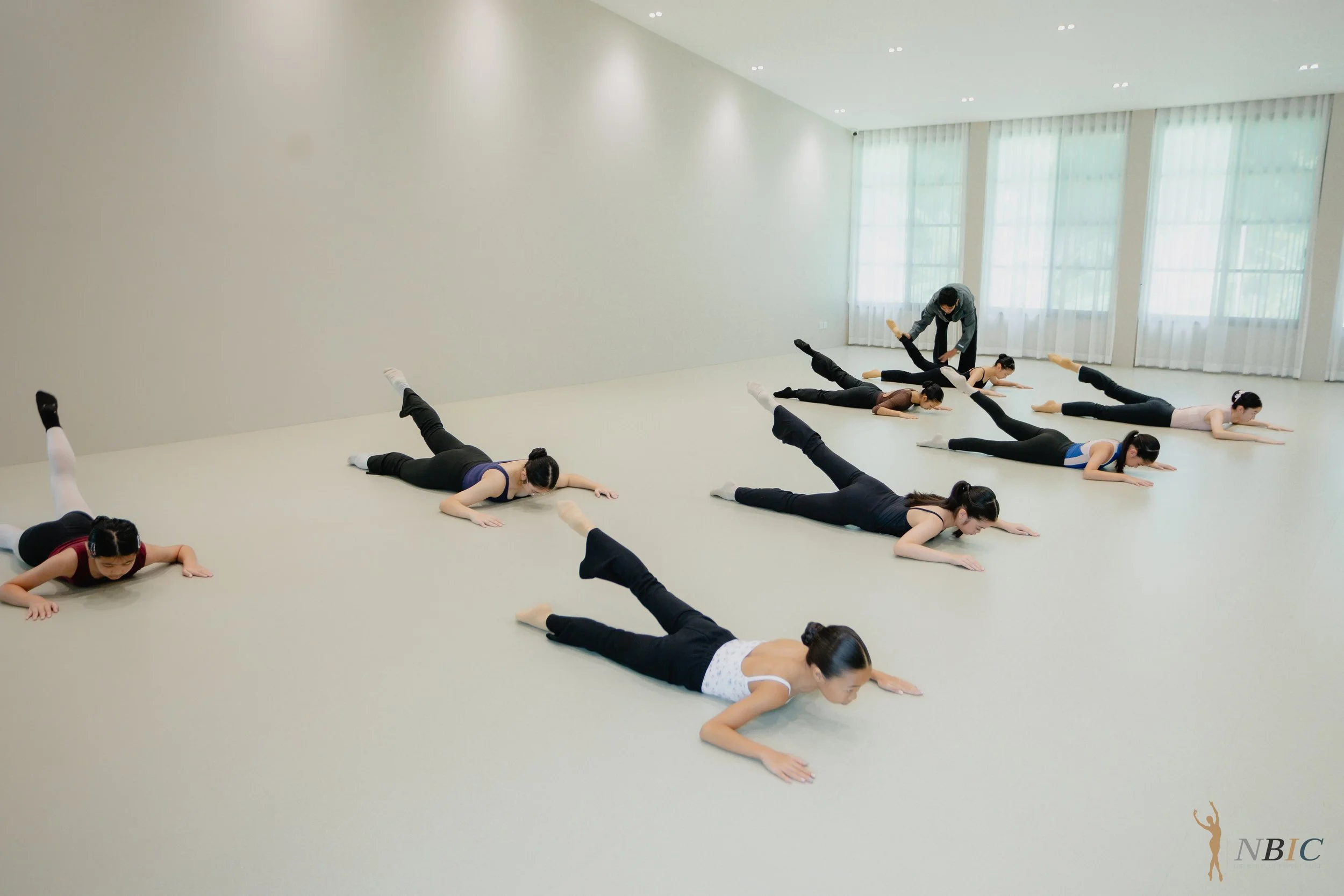 A group of ballet dancers practicing in a spacious, brightly lit dance studio with high ceilings and large windows. They are lying on the floor in a split position, stretching with their arms extended forward, guided by an instructor.