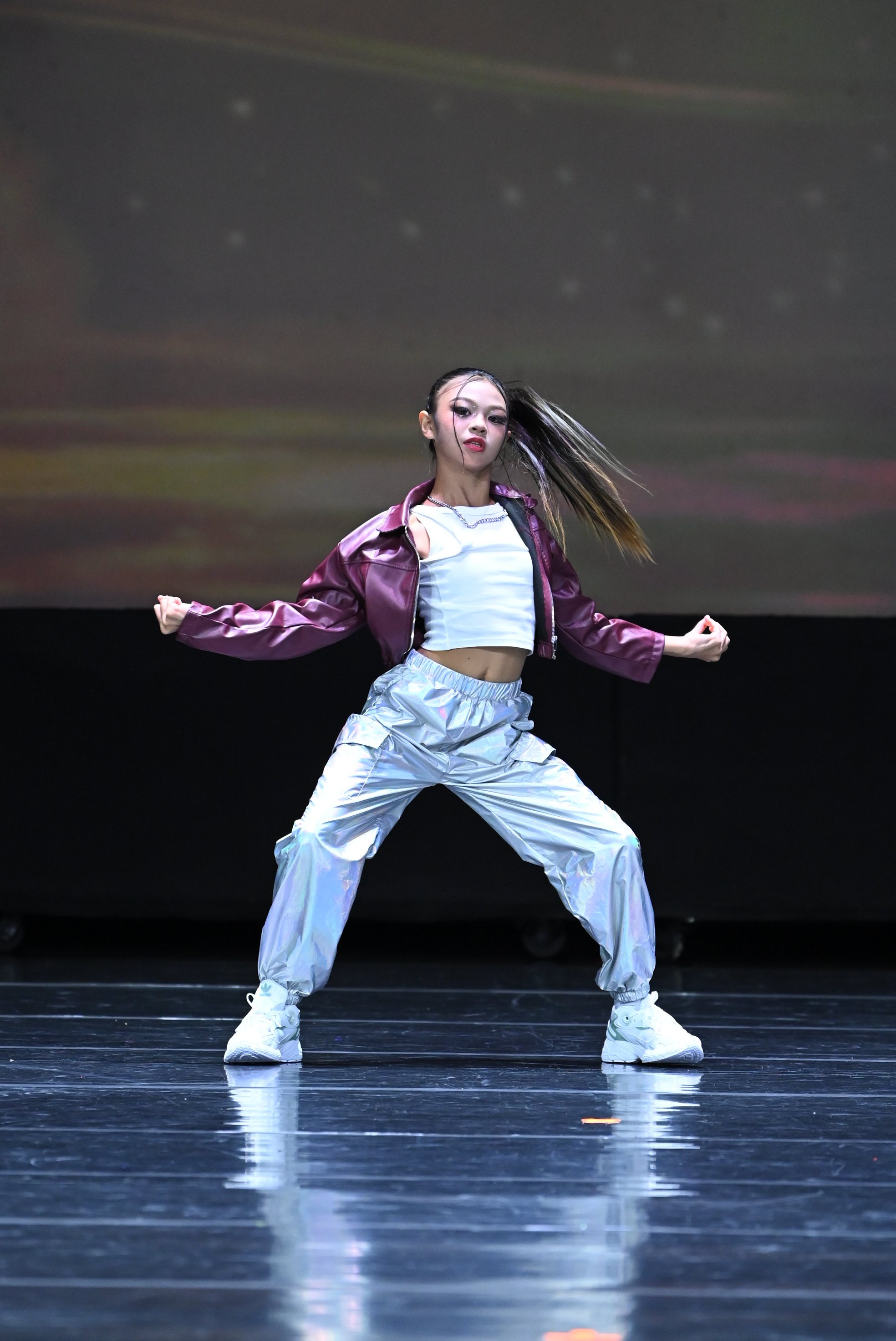 A female dancer performing on stage, wearing metallic silver pants, a white crop top, and a maroon jacket, with her hair flowing and a focused expression.