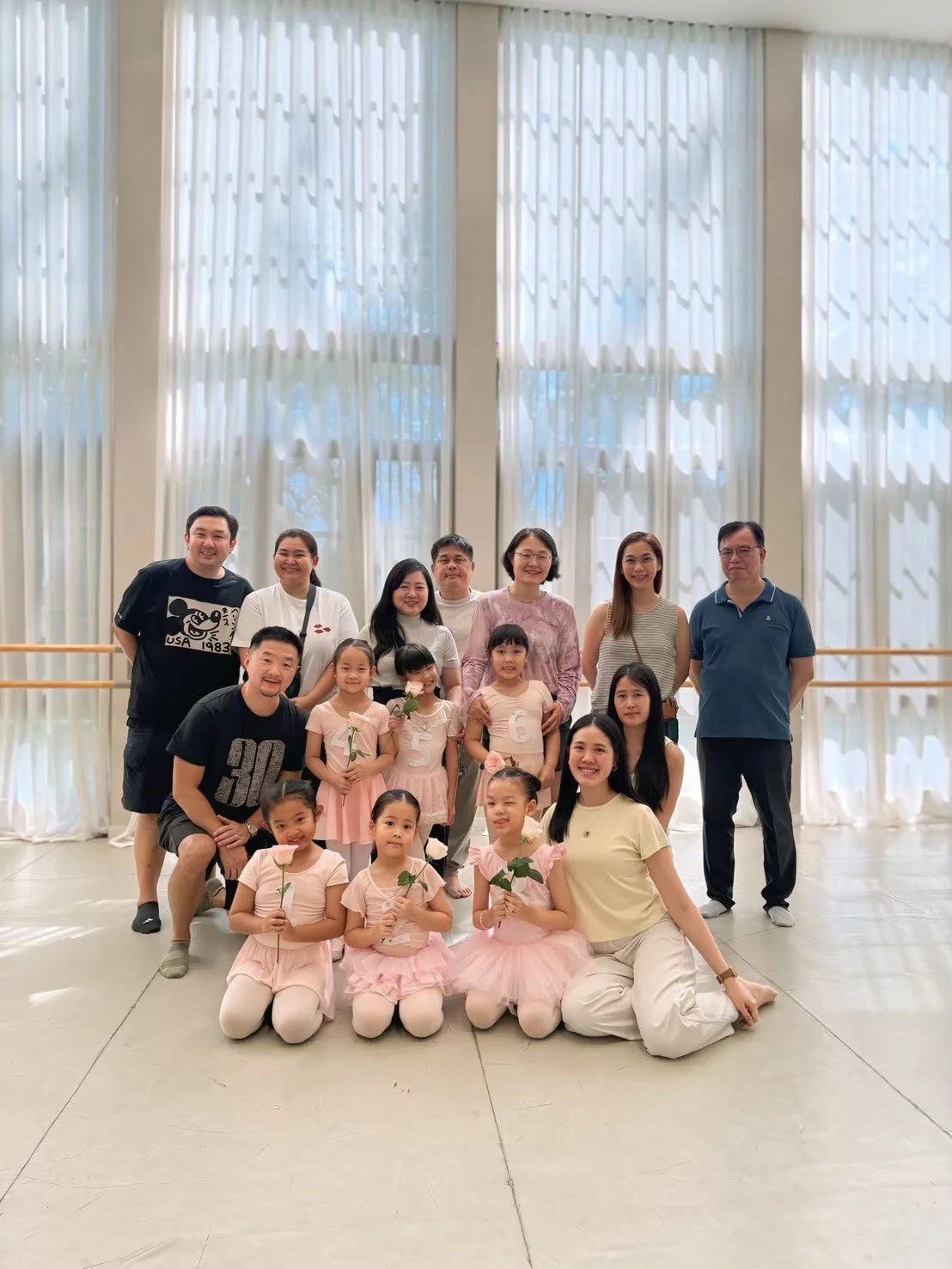 Group photo of children and adults standing and sitting in a spacious room with large windows and sheer curtains, some children holding flowers, celebrating a special occasion.