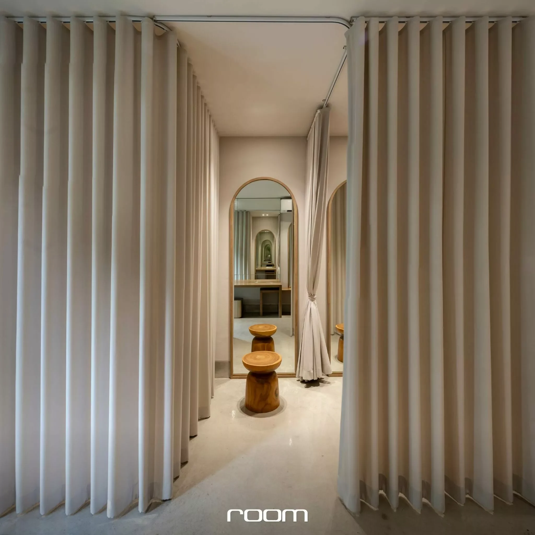 View of a fitting room with beige curtains, a mirror on the wall, and wooden stools on a light-colored floor.