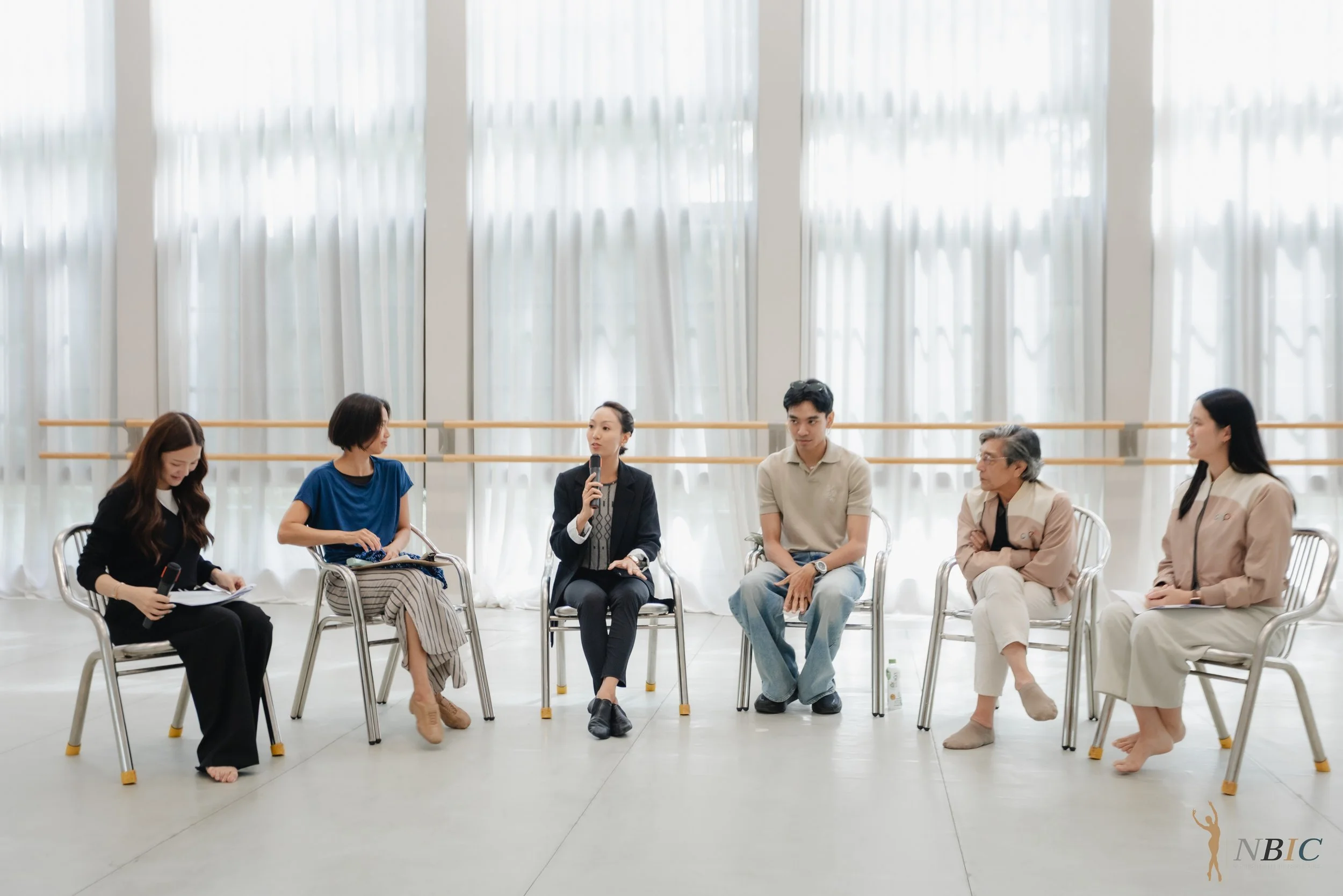 A group of six people sitting in chairs in a bright room with large, sheer window curtains. They are participating in a discussion or panel, with one woman holding a microphone and speaking. Others are listening attentively, some with microphones and