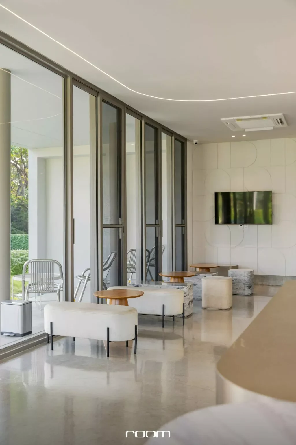 Modern interior lounge area with white and beige furniture, small wooden tables, a wall-mounted flat-screen TV, large glass windows, and outdoor greenery visible through the windows.