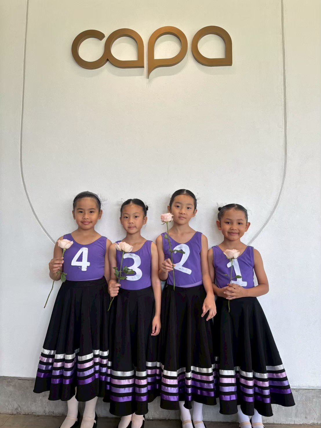 Four young girls dressed in purple and black costumes, holding pink roses, standing in front of a white wall with the word 'capa' on it.