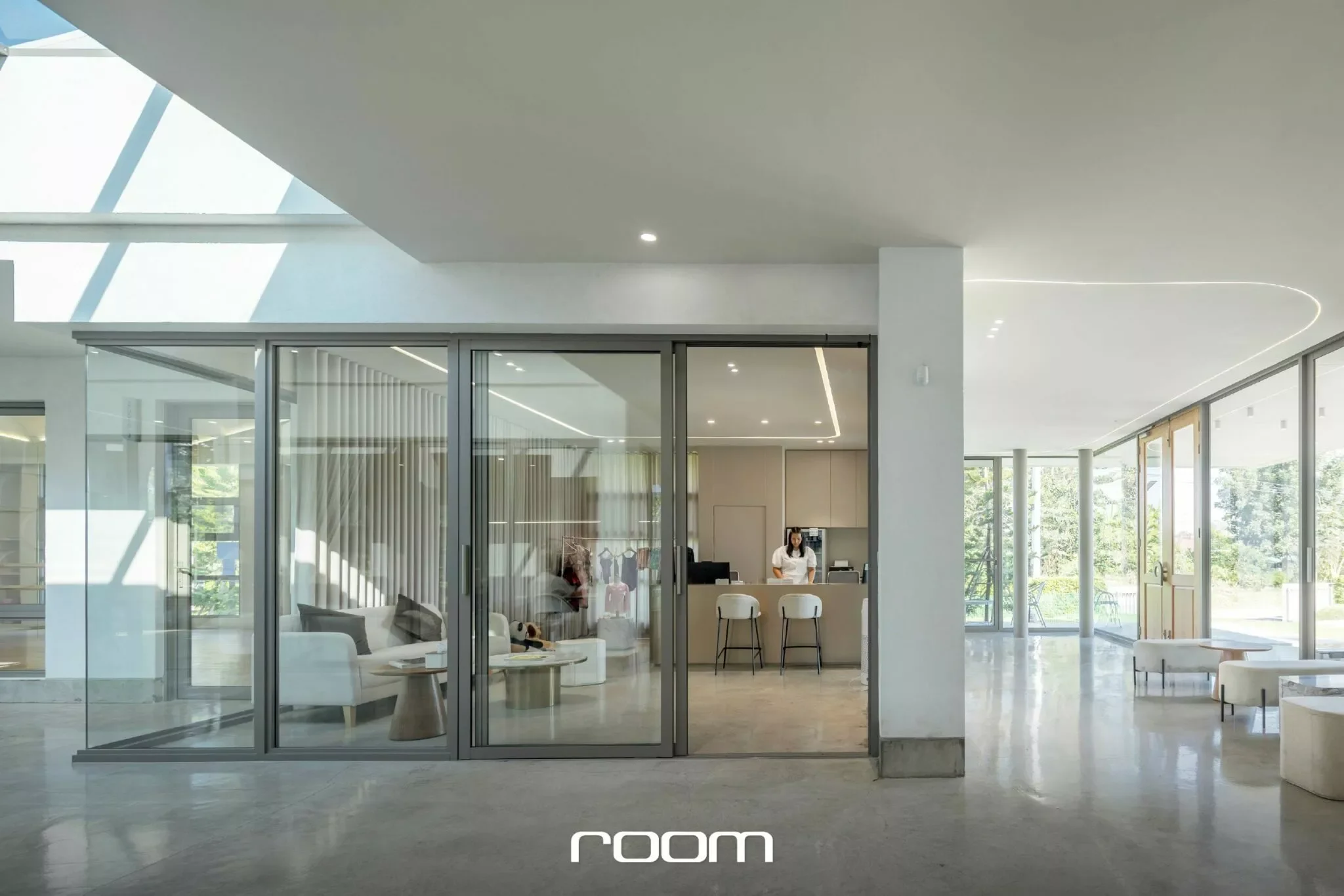 Modern interior with glass walls, a white sofa, and a reception area with a woman standing behind a counter, sunlight streaming through large windows.