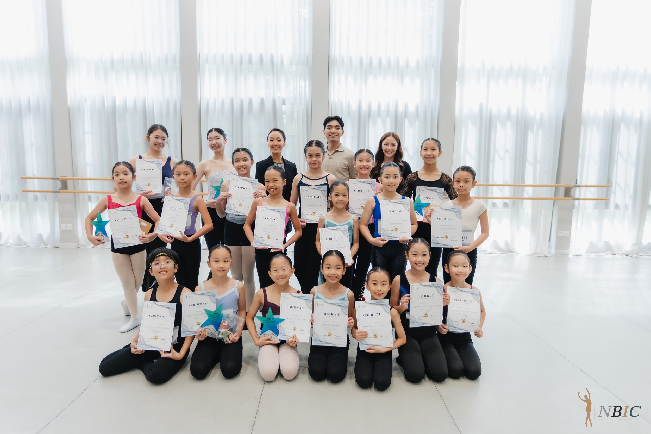 Group of young ballet dancers holding certificates and awards, posing for a photo in a dance studio with large windows and ballet barres, along with adult instructors.