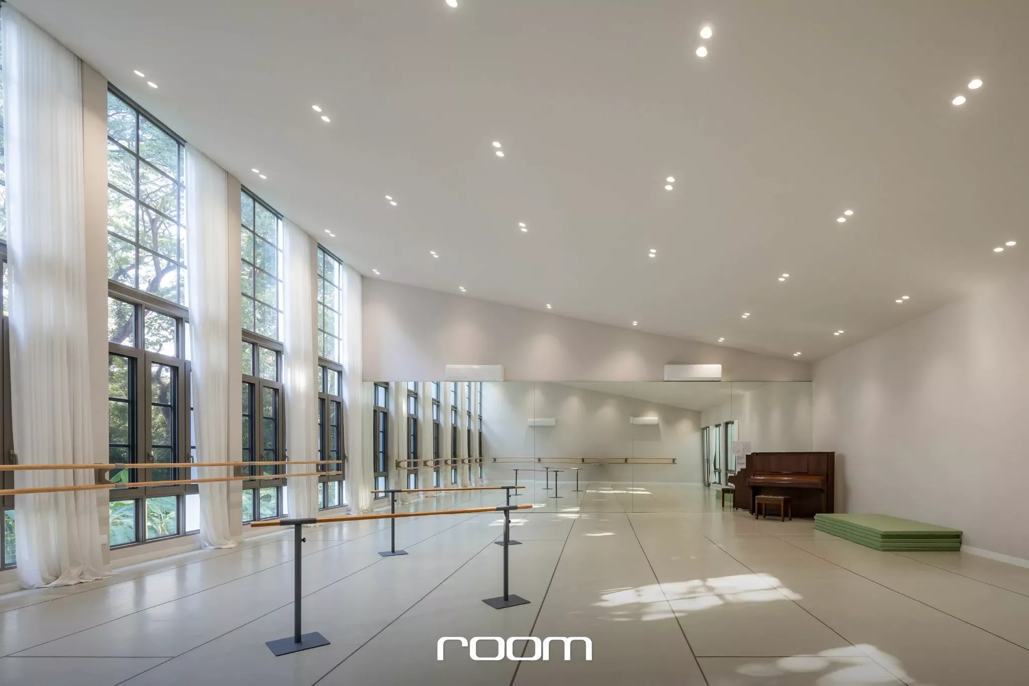 A spacious dance studio with large windows, white curtains, and a wooden bar along the wall. The room is well-lit with ceiling lights, and a piano is visible in the corner.