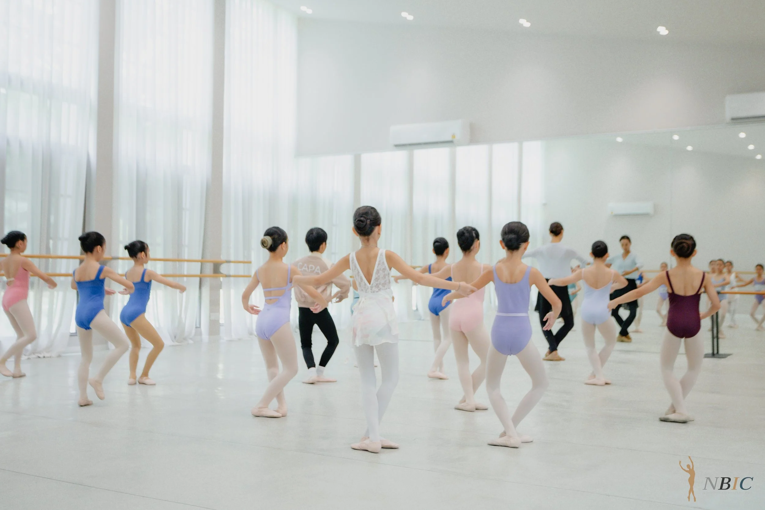 Ballet dancers practicing or rehearsing in a brightly lit studio with large windows and mirrors, wearing leotards and tights.