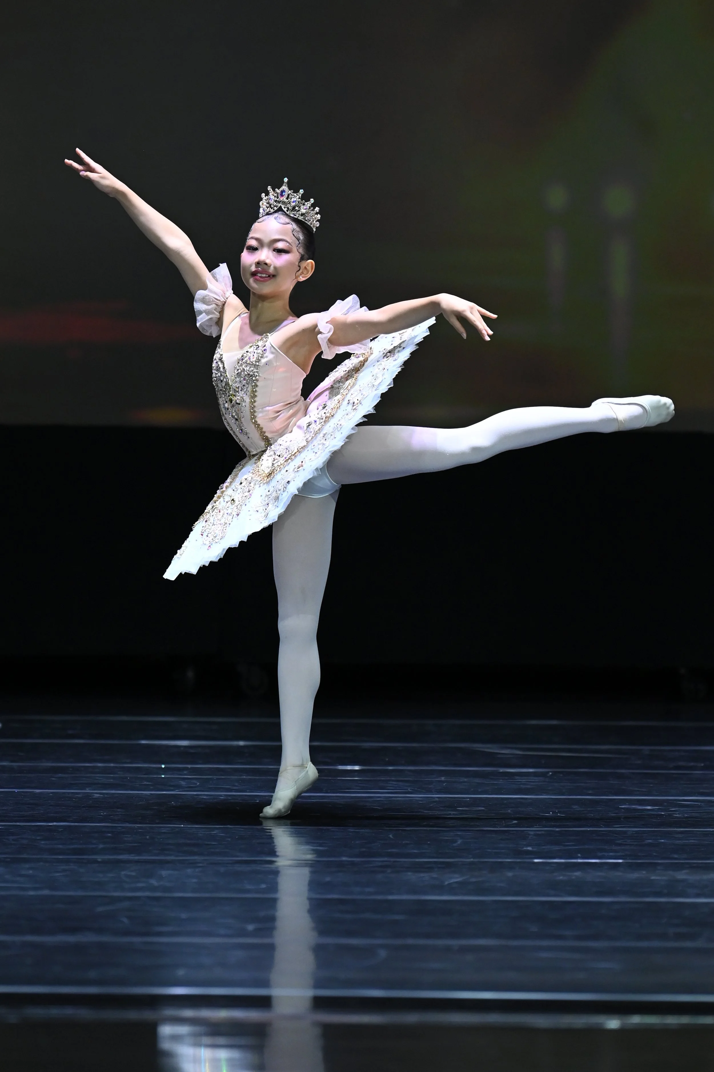 A young ballet dancer performing a pose on stage, wearing a tiara and a detailed tutu with embellishments.
