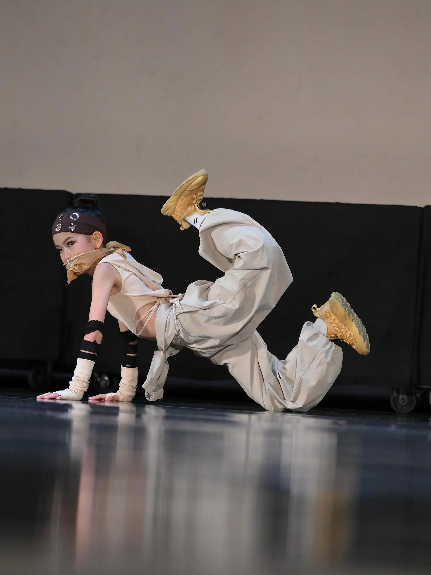 A young performer in a beige outfit, with a headband and makeup, is on the floor in a dance pose with hands and knees on the stage, next to another performer in a baggy beige outfit and yellow shoes, who's also in a dance pose.