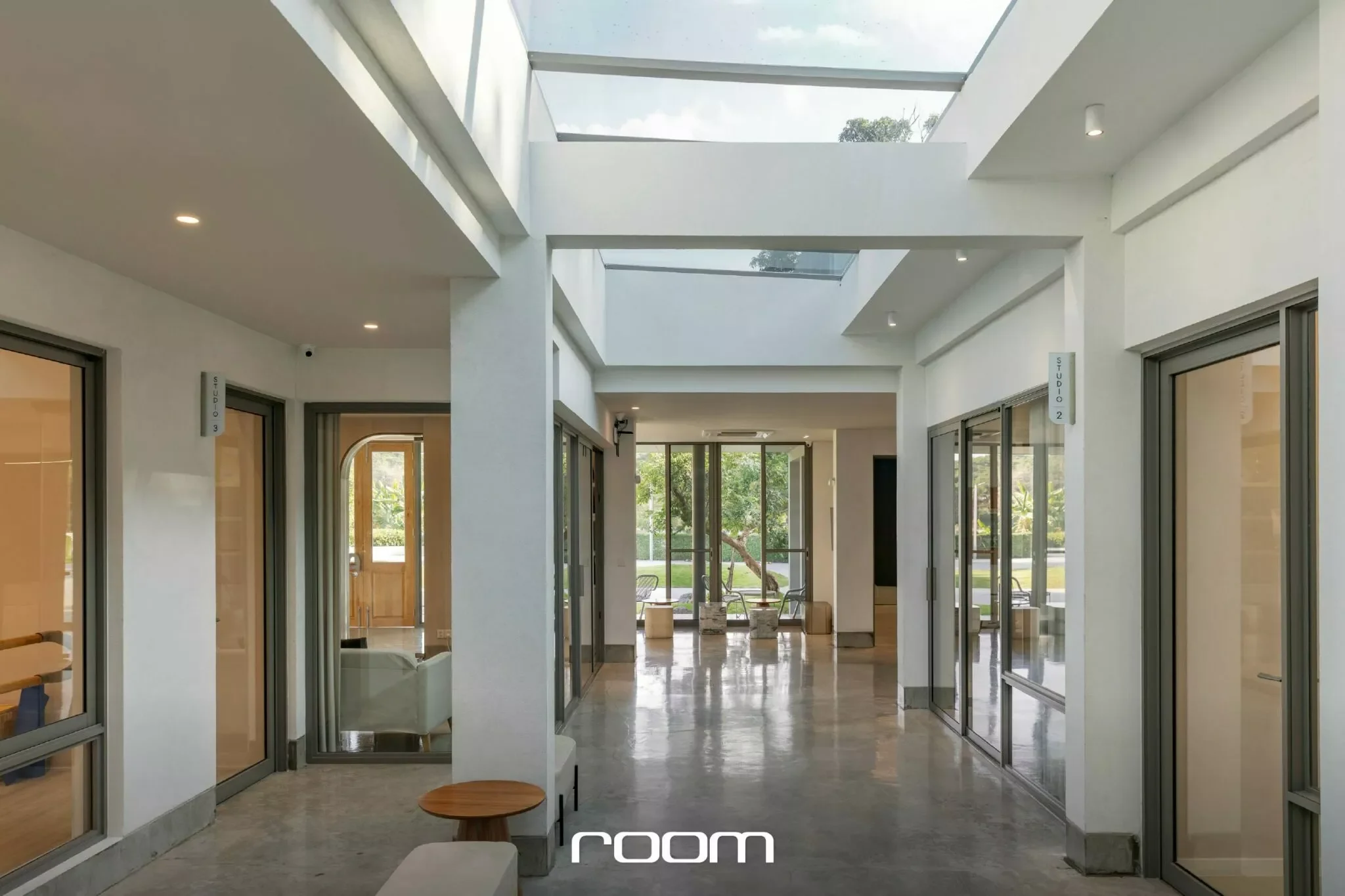 Modern interior hallway with large glass windows, a skylight ceiling, and outdoor greenery visible through the windows and glass doors.