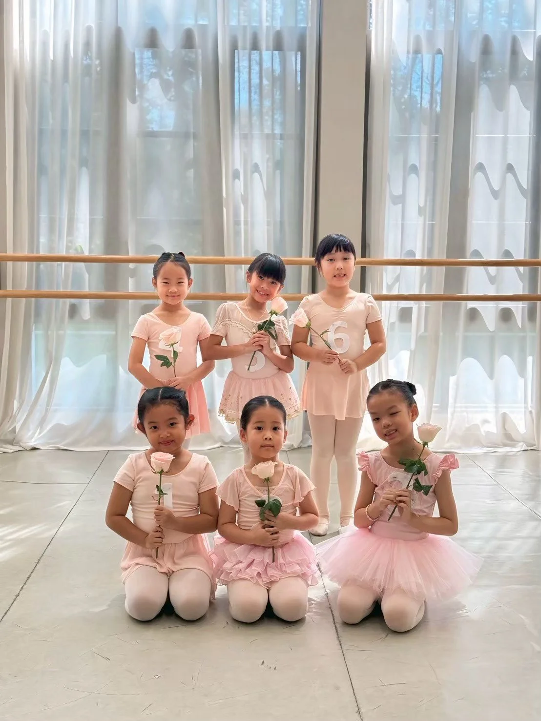 Seven young girls dressed in ballet costumes, holding pink roses, posing in a ballet studio with large windows and sheer curtains.