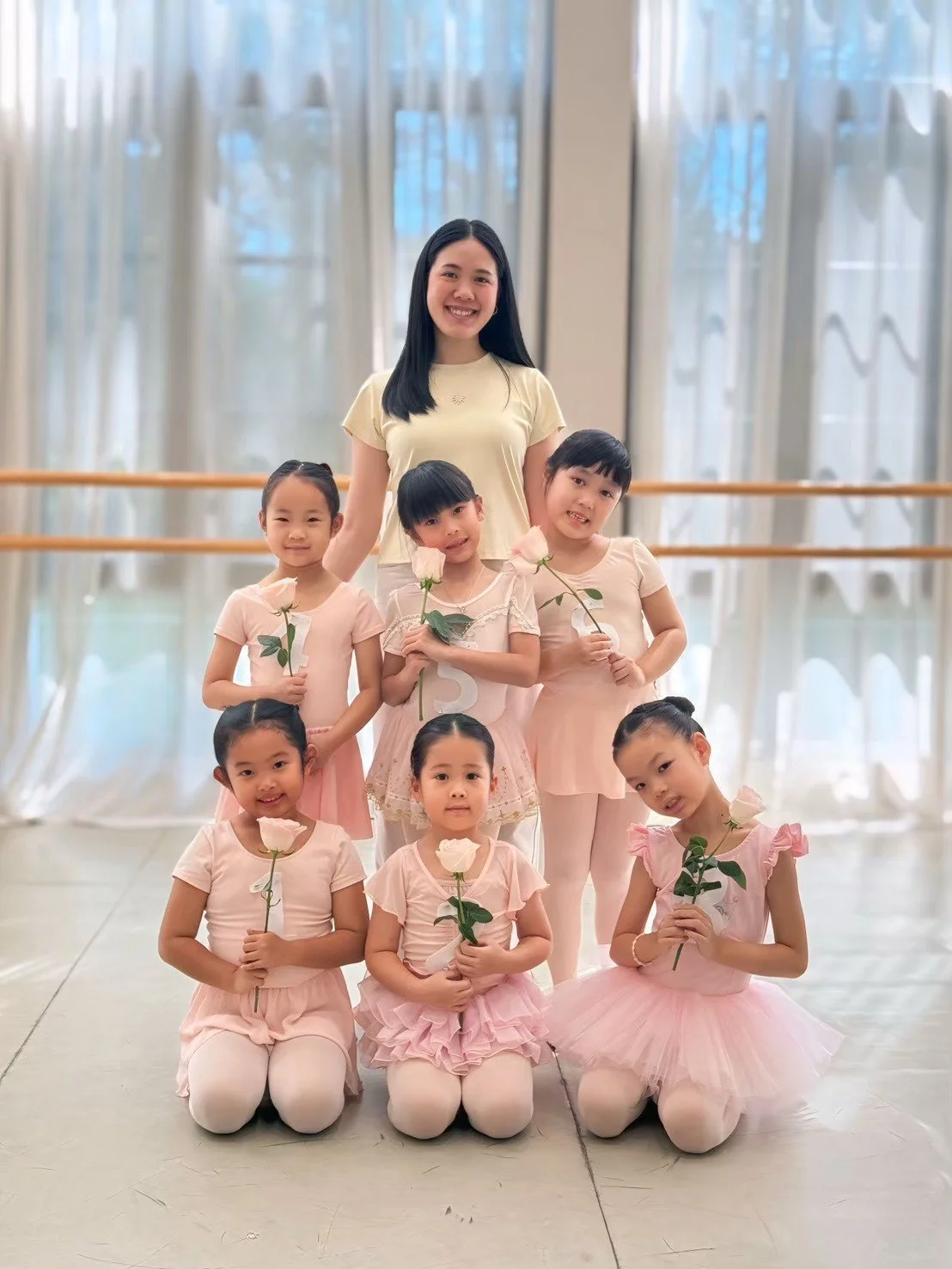 A group of young girls and a woman, possibly a dance instructor, standing in a dance studio with sheer curtains and ballet bars in the background. The girls are holding roses and wearing light pink and cream dance costumes. The woman is smiling and w