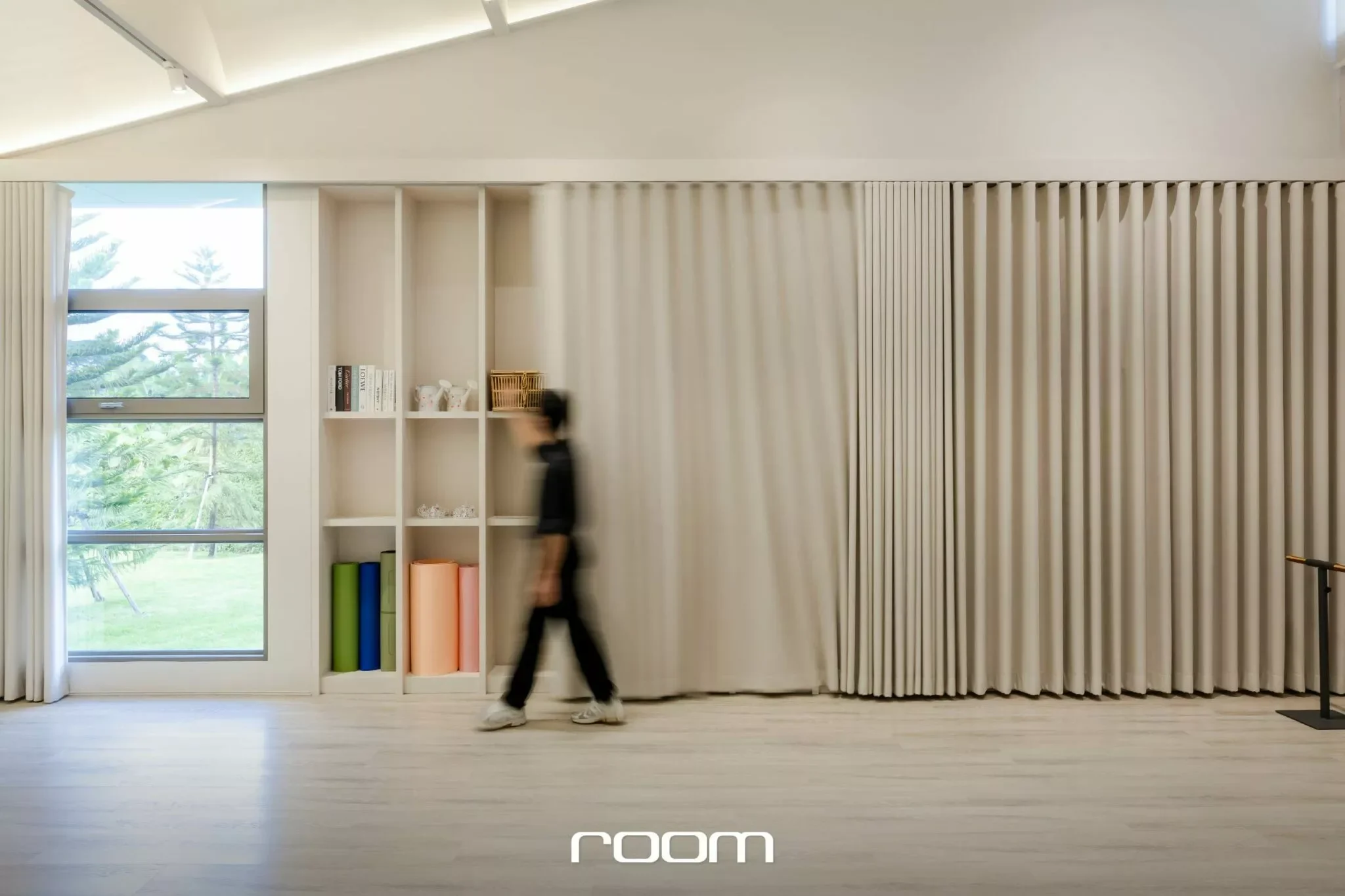 A person walking in front of a modern interior with large windows, beige curtains, a white bookshelf with books and decor, and light wooden flooring.