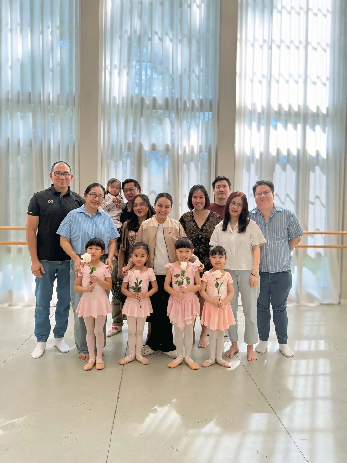 A group photo of adults and children standing inside a bright room with large windows and white curtains. The children in front are holding white roses, and the group is smiling for the camera.