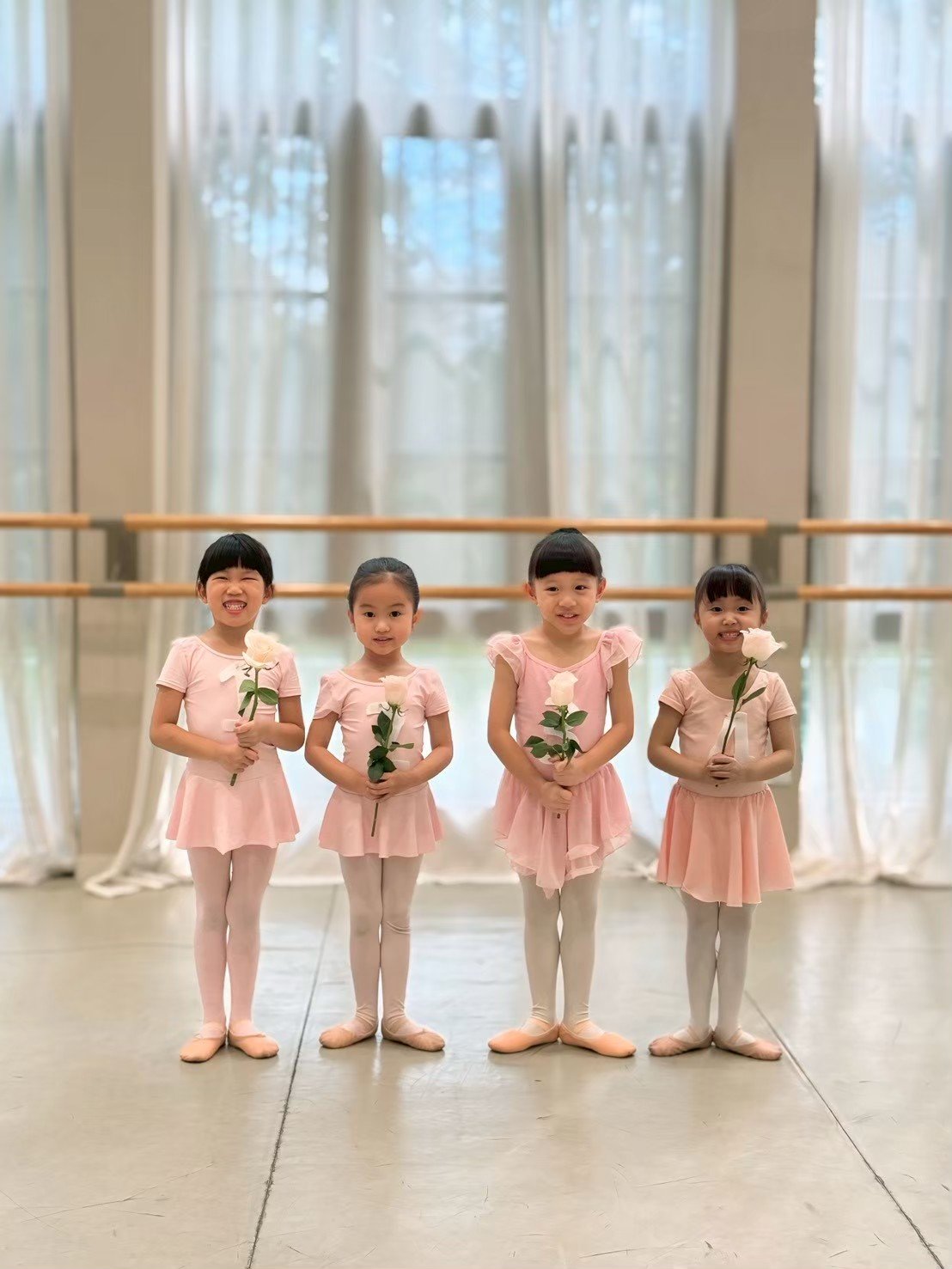 Four young girls dressed in pink ballet outfits holding white roses pose in a dance studio with large windows and curtains.