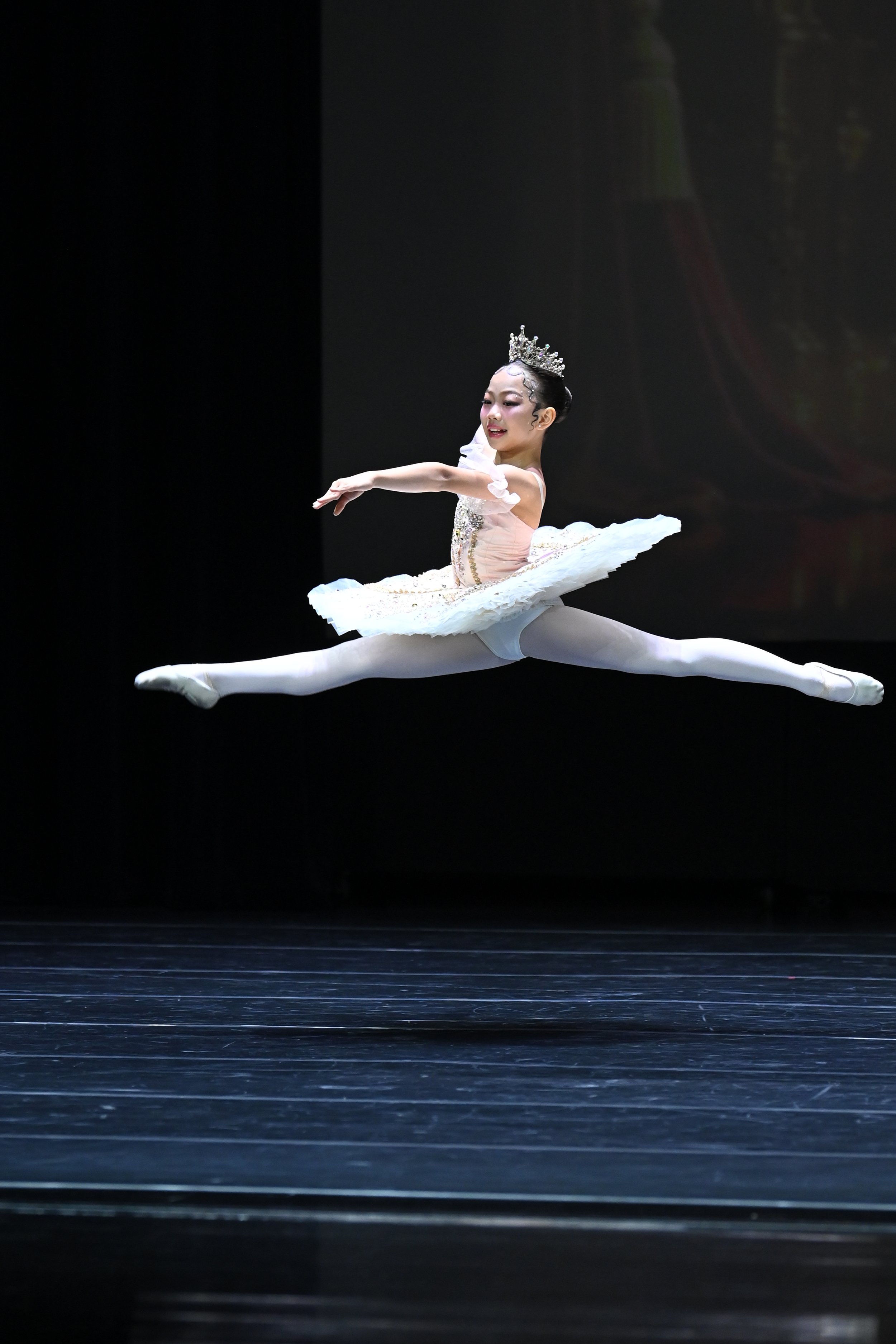 Young girl in ballet costume performing a split leap on stage, wearing a tiara and white tights, with a dark background.