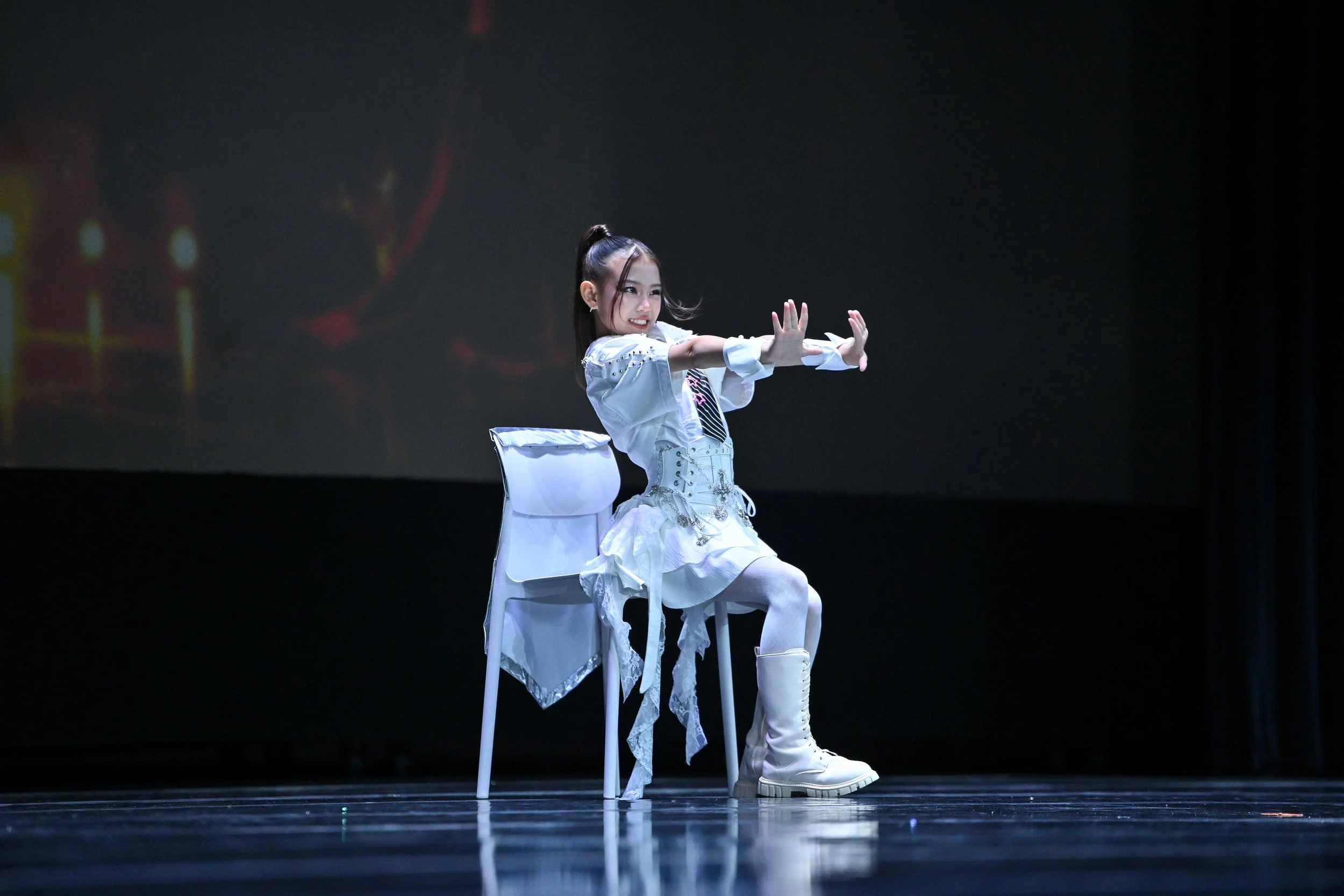 A young woman dressed in a white outfit performing a dance move on stage, sitting on a chair with her arms extended forward and smiling.