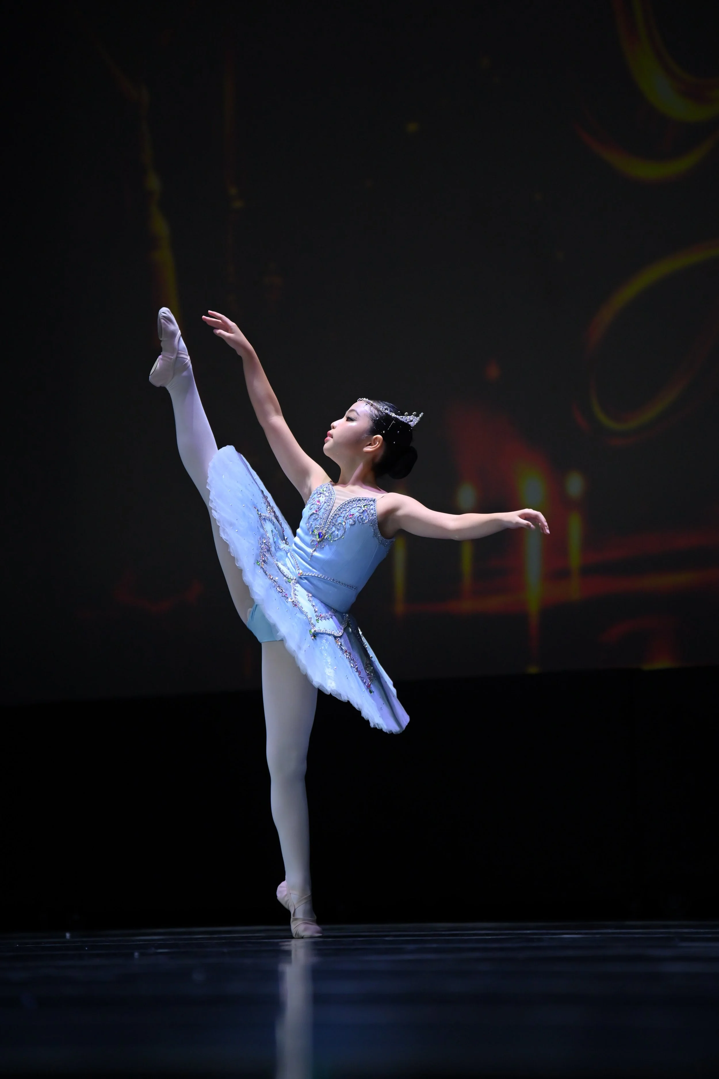 Ballet dancer performing a high kick on stage, wearing a light blue tutu, with a dark background.