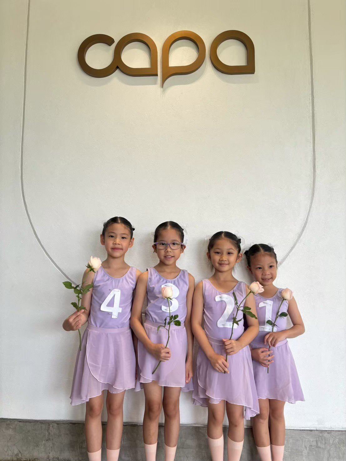 Four young girls in purple dresses holding white roses with number labels, standing in front of a wall with a large 'capa' sign, likely at a dance or graduation event.